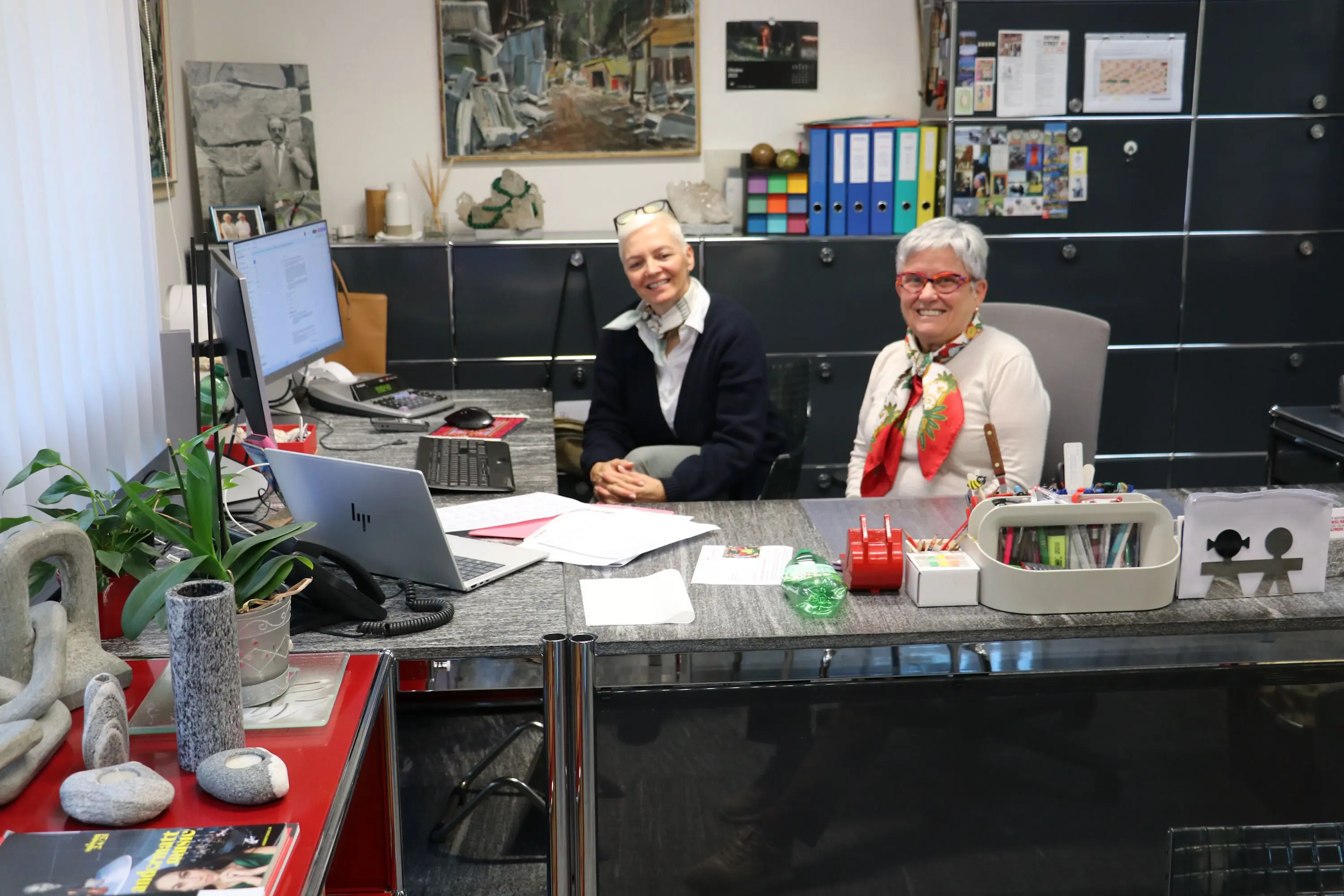 Due donne sorridenti sedute alla scrivania di un ufficio. Una di loro indossa occhiali e ha i capelli corti, mentre l'altra ha i capelli grigi e indossa un maglione chiaro. Sul tavolo ci sono un computer, documenti e materiali da ufficio. Sullo sfondo si vedono opere d'arte e armadietti