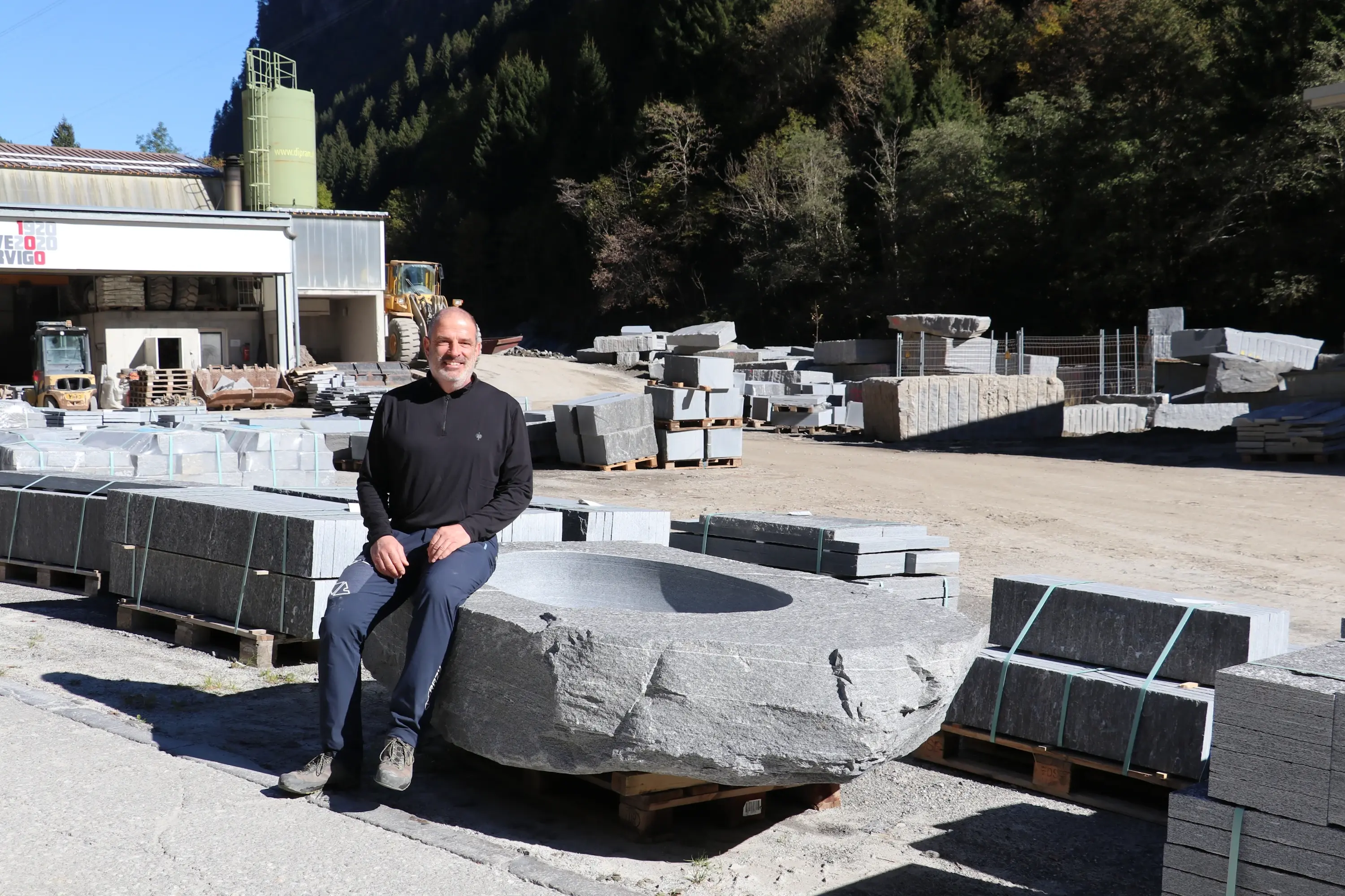 Un uomo sorridente si siede su una grande pietra al centro di un cantiere di lavorazione della pietra, circondato da blocchi di granito e attrezzature. Sullo sfondo si vedono alberi e una struttura industriale.