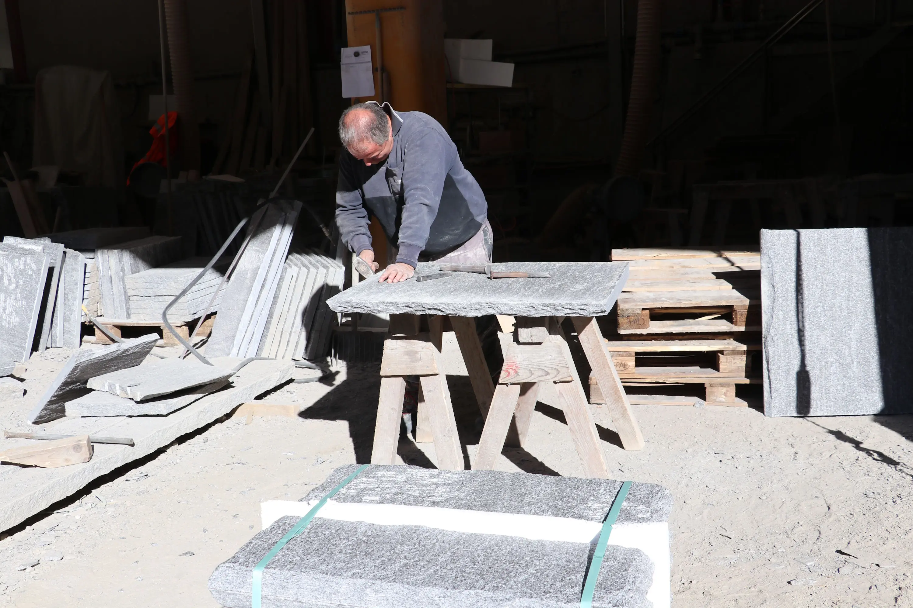 Un uomo lavora su un pezzo di pietra in un laboratorio. È circondato da blocchi di pietra e attrezzi, mentre un fascio di luce illumina l'area.