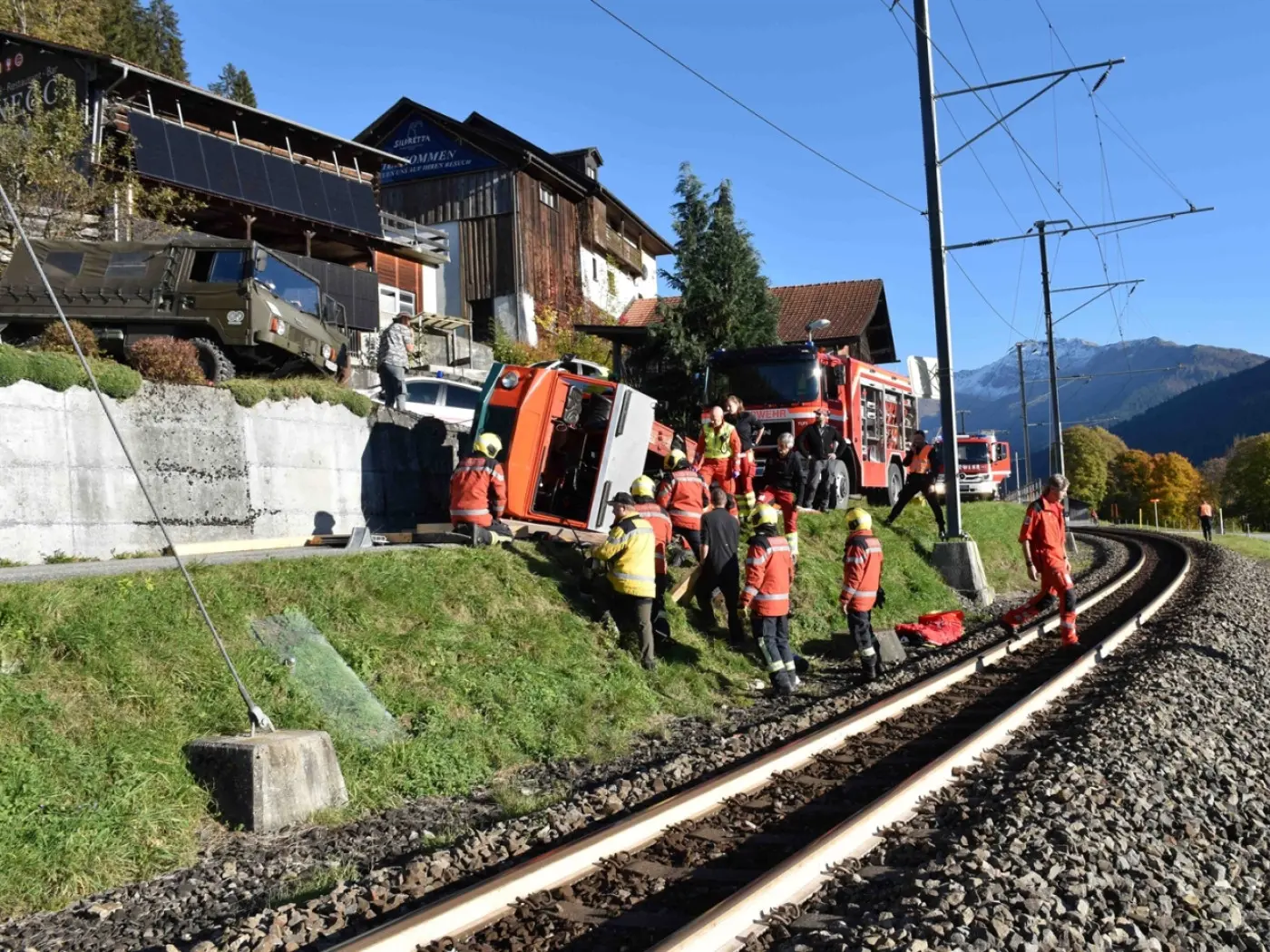 Intervento di emergenza con personale dei vigili del fuoco e soccorritori intorno a un camion ribaltato vicino ai binari del treno, in un'area montana con case e alberi sullo sfondo.