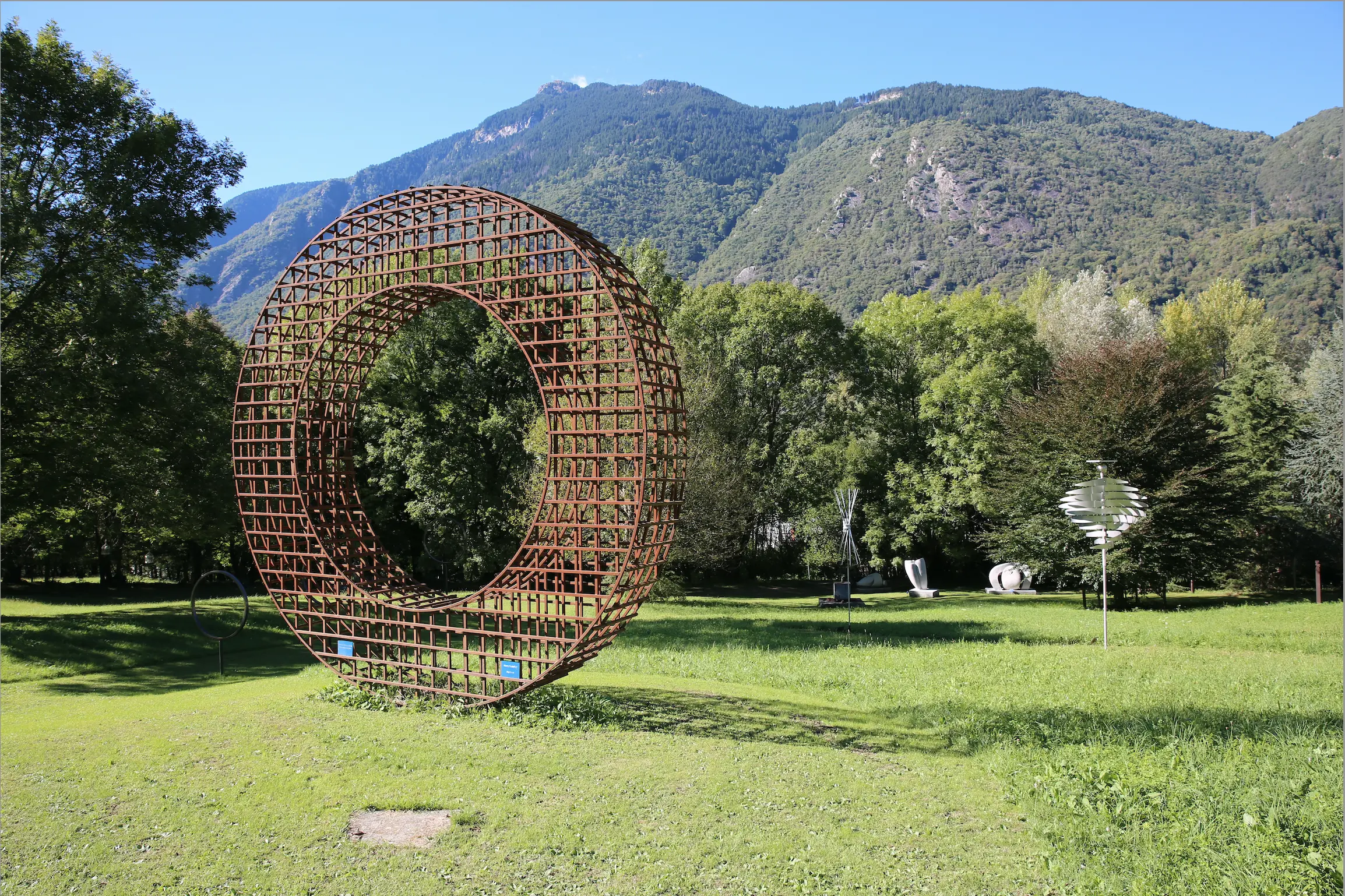Una grande scultura a forma di anello in metallo, posizionata in un parco verde circondato da alberi e montagne sullo sfondo.
