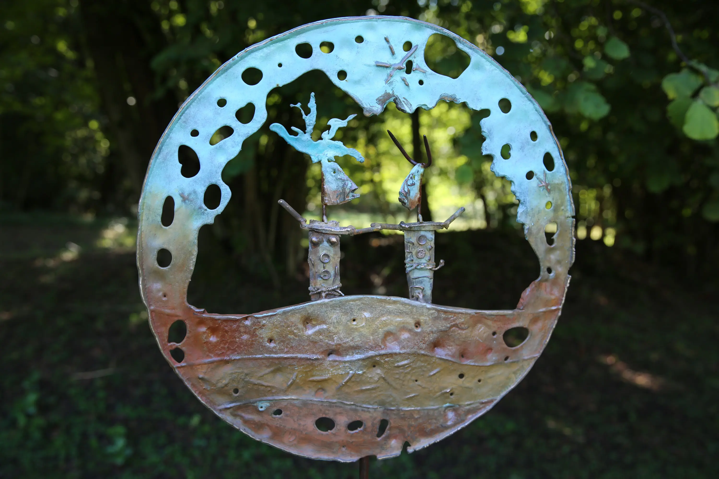 Scultura in metallo rappresentante due figure stilizzate in un paesaggio circolare, con dettagli di alberi e un cielo blu.