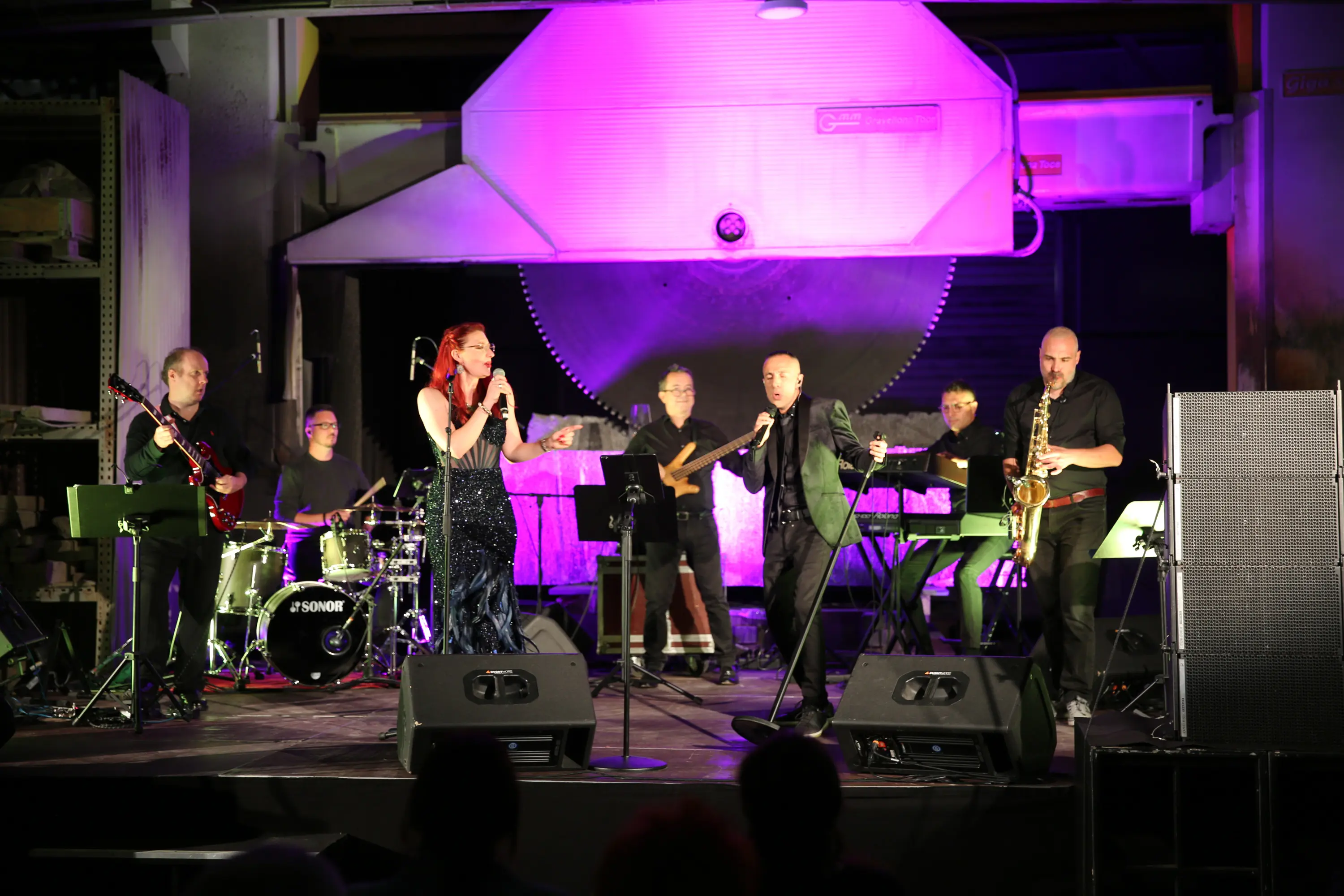 Un concerto dal vivo con otto musicisti sul palco. Una cantante è al centro, mentre il sax e la batteria accompagnano. Luce viola illumina l'area, creando un'atmosfera suggestiva.