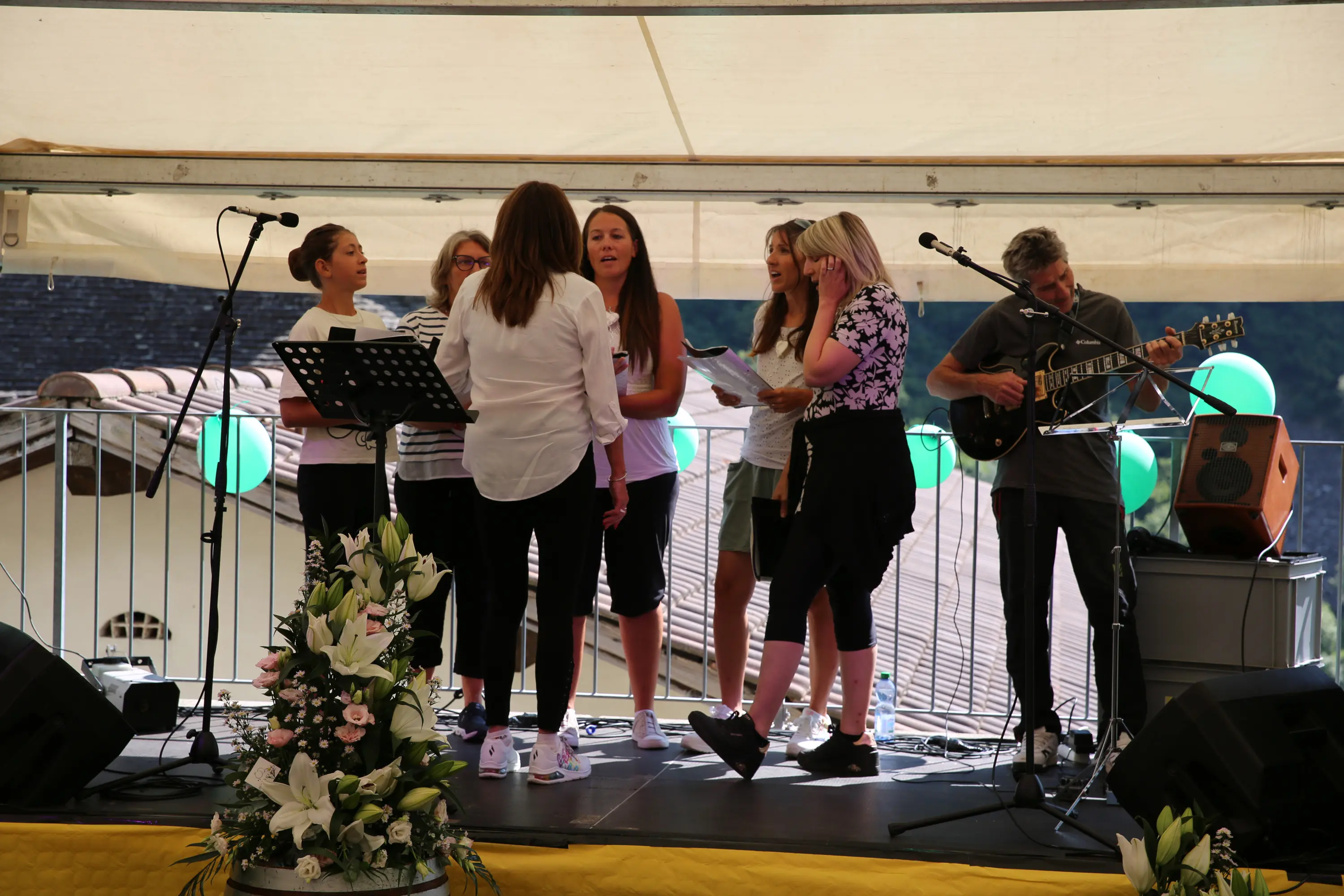 Un gruppo di sei cantanti è in scena durante una performance. Alcune ragazze indossano magliette leggere e si concentrano sulle loro parti. Un musicista suona la chitarra accanto a loro, mentre un microfono e uno spartito sono visibili sul palco. Sullo sfondo, si intravedono decorazioni verdi e un tetto di un edificio.