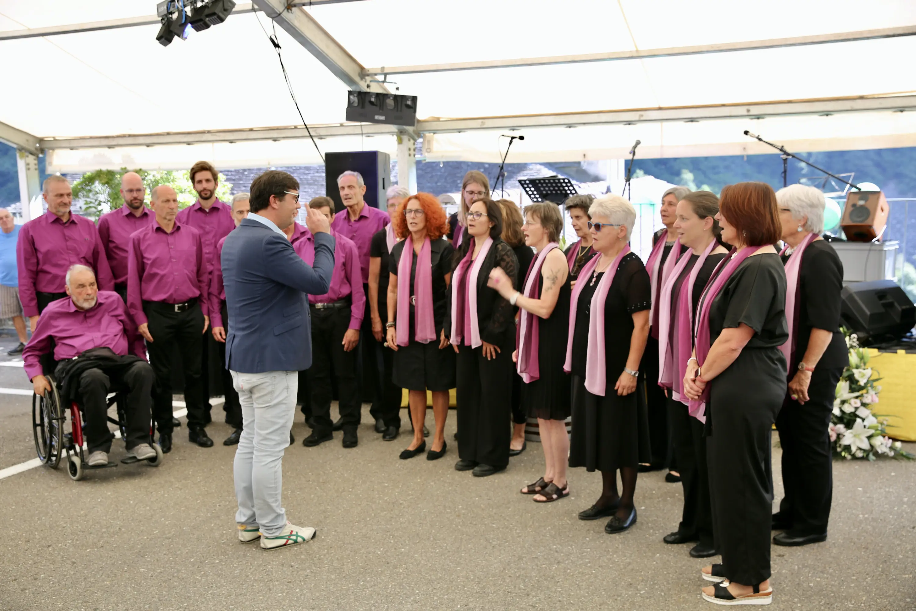 Un gruppo di cantanti in abiti neri e camicie fucsia, con alcune sciarpe bianche, si esibisce sotto un tendone. Un direttore di coro sta orchestrando la performance. In seconda fila, una persona è seduta su una sedia a rotelle. Sullo sfondo si vedono strumenti musicali.
