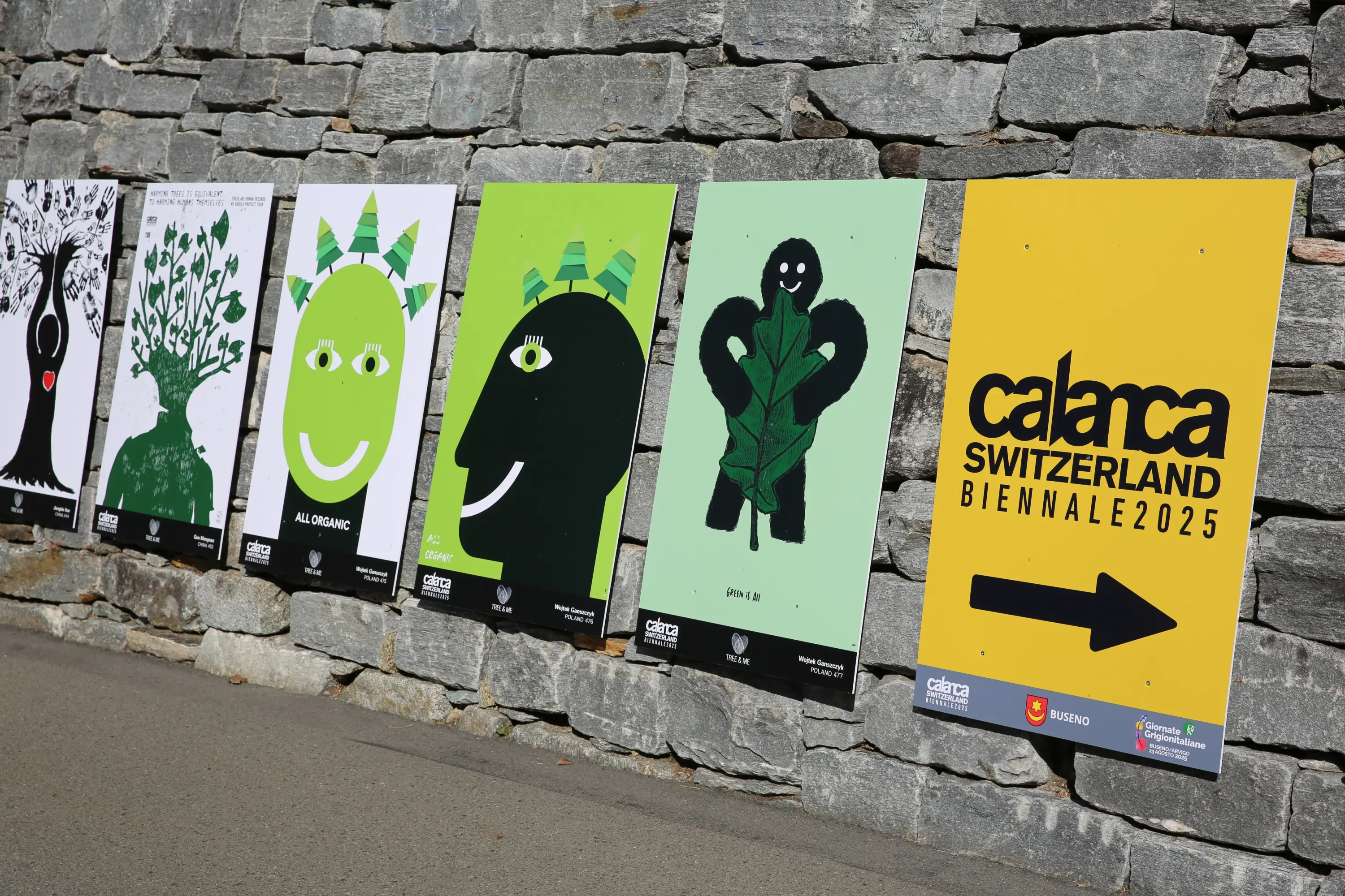 Varie illustrazioni colorate affisse su un muro di pietra, in vista laterale. Le opere presentano volti stilizzati e motivi naturali, con un grande pannello giallo che indica la Biennale di Calanca in Svizzera nel 2025.
