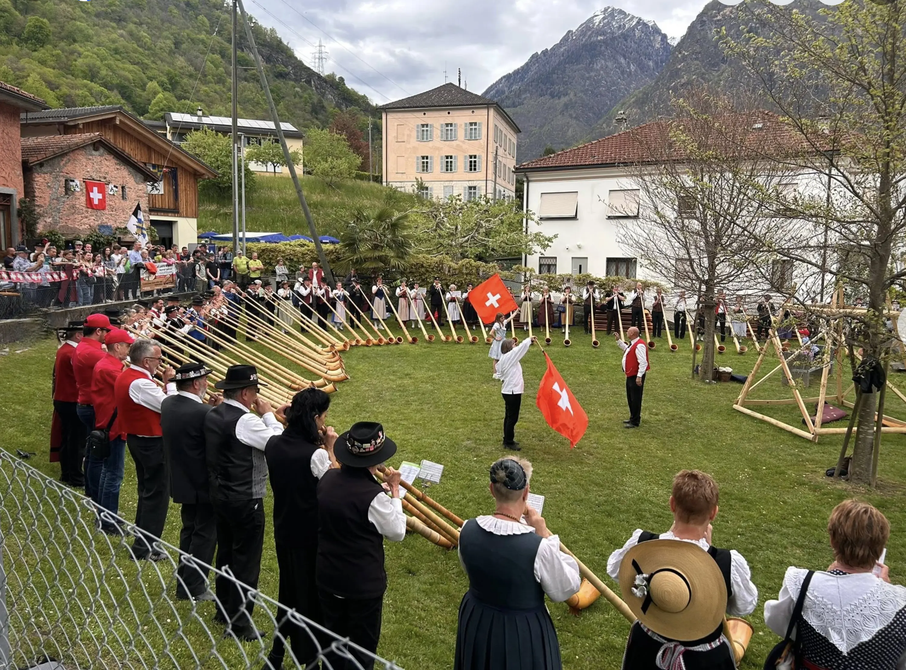 Gruppo di persone in costume tradizionale svizzero si riunisce in un prato, mentre suonano gli alpenhorn e sventolano bandiere svizzere. Sullo sfondo, un pubblico assiste all'evento.