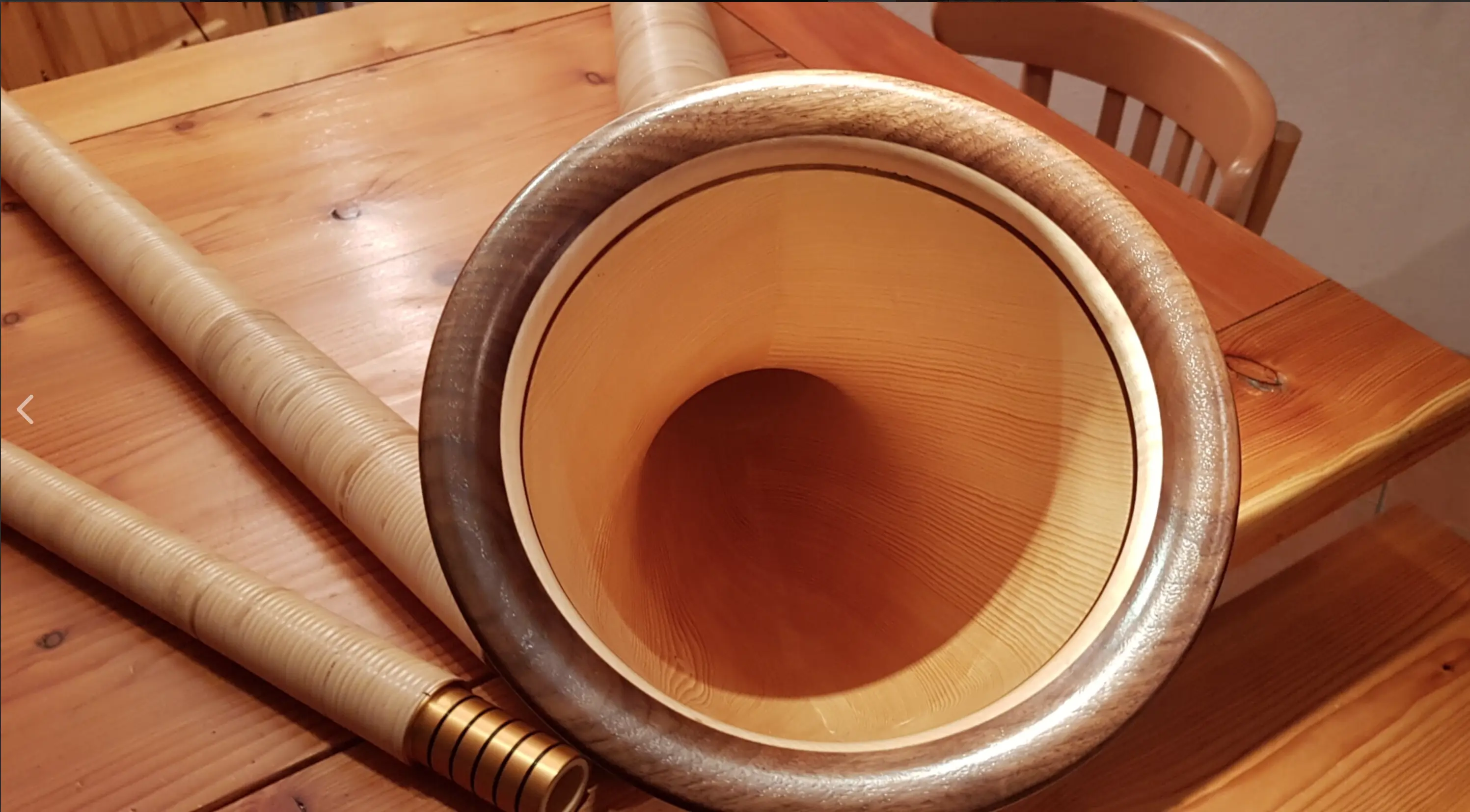 Un corno in legno con una bocca ampia, posato su un tavolo di legno chiaro. Dietro di esso, si vedono due tubi di legno di diverse dimensioni.