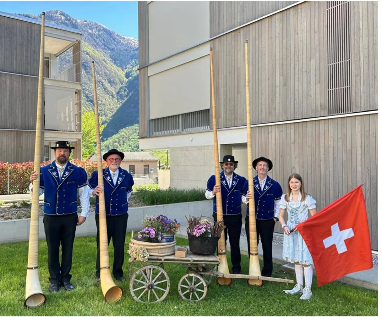 Gruppo di uomini in abiti tradizionali svizzeri, ciascuno con un corno delle Alpi, accanto a una ragazza che tiene una bandiera svizzera. Dietro di loro, un carretto con fiori e un paesaggio montano sullo sfondo.