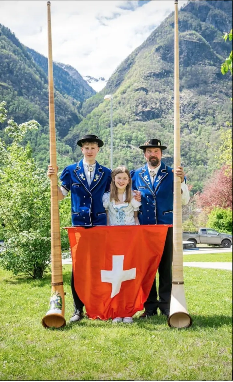 Tre persone posano accanto a grandi corni alpini, indossando costumi tradizionali svizzeri. Due uomini a sinistra e una ragazza al centro tengono una bandiera svizzera rossa con una croce bianca in mezzo. Sullo sfondo si vedono montagne verdi.