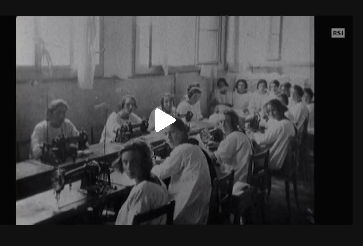 Un laboratorio di sartoria con diverse donne sedute a macchine da cucire. Le operatrici indossano camici bianchi e lavorano in un ambiente luminoso e spazioso. Alcune donne guardano verso la telecamera.