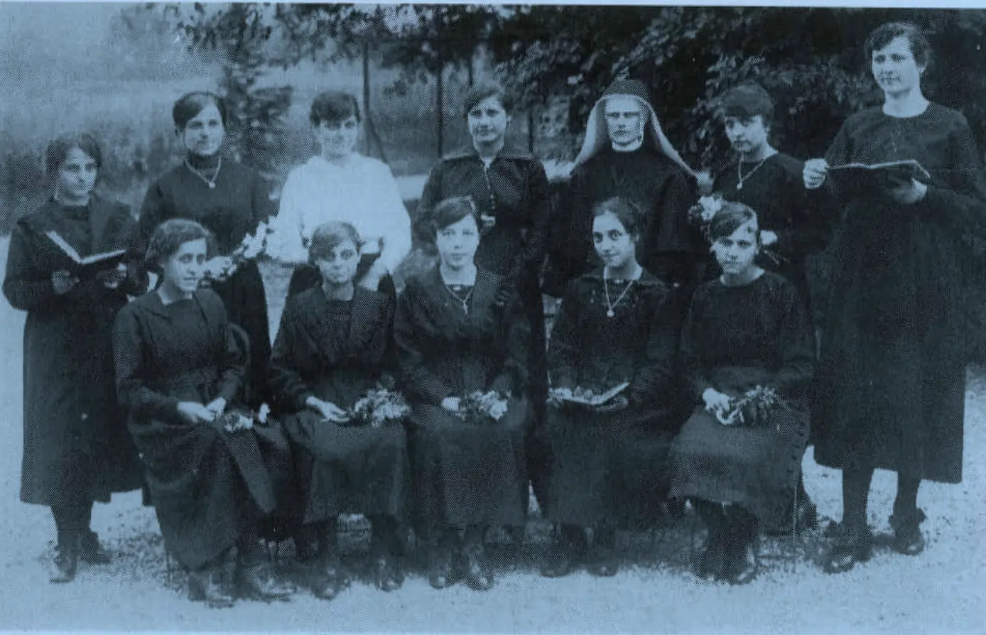 Gruppo di dodici donne in abiti scuri, molte con fiori in mano, posano all'aperto in un giardino. Alcune portano crocifissi al collo e una suora è presente tra di loro. L'atmosfera è formale e nostalgica.