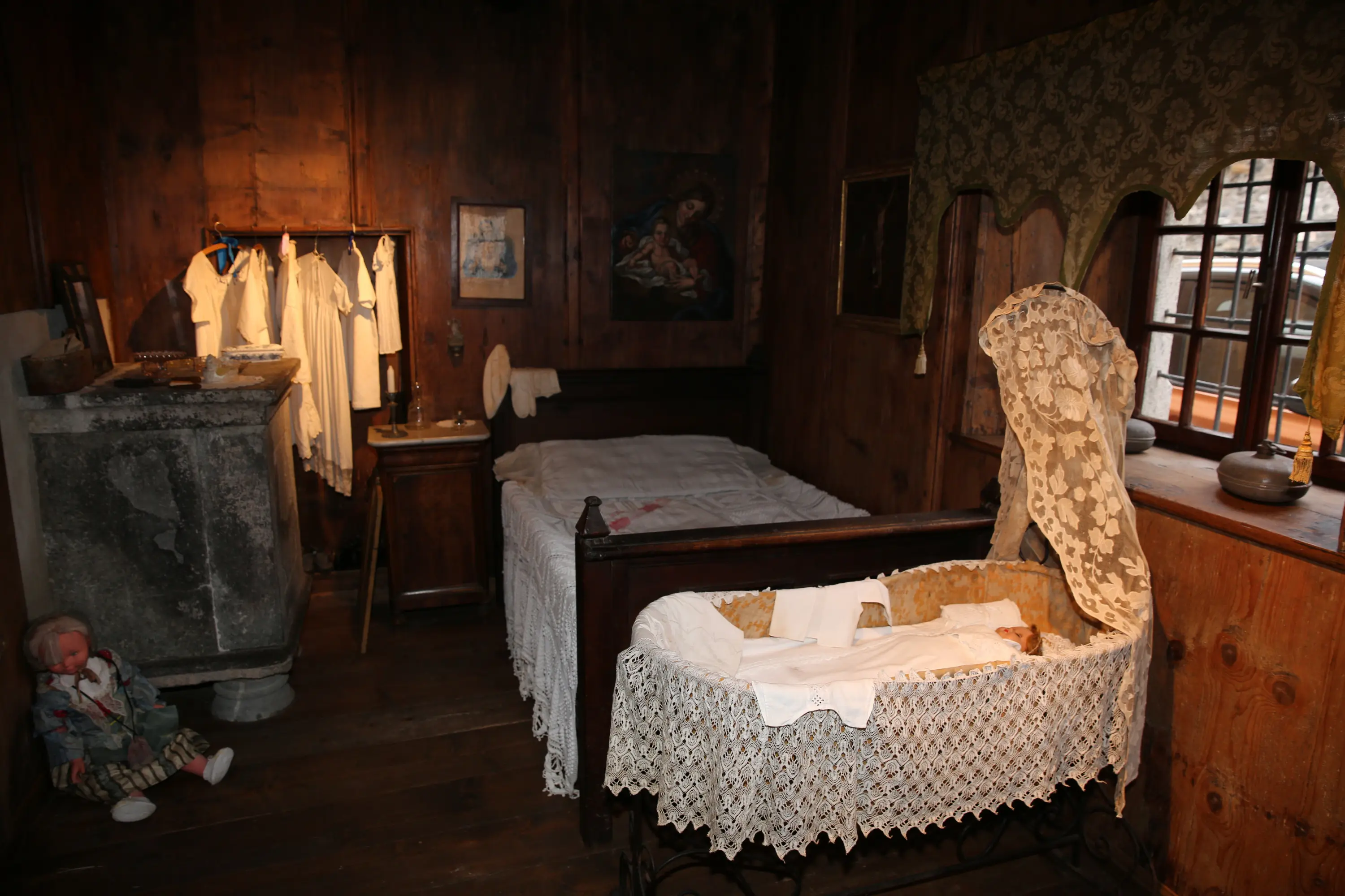 Una stanza rustica con pareti di legno, un letto semplice coperto da una biancheria bianca, un fasciatoio con panni bianchi e un culla decorata con pizzo. Un vecchio bambolotto è appoggiato a terra accanto a una finestra con tende a motivo.