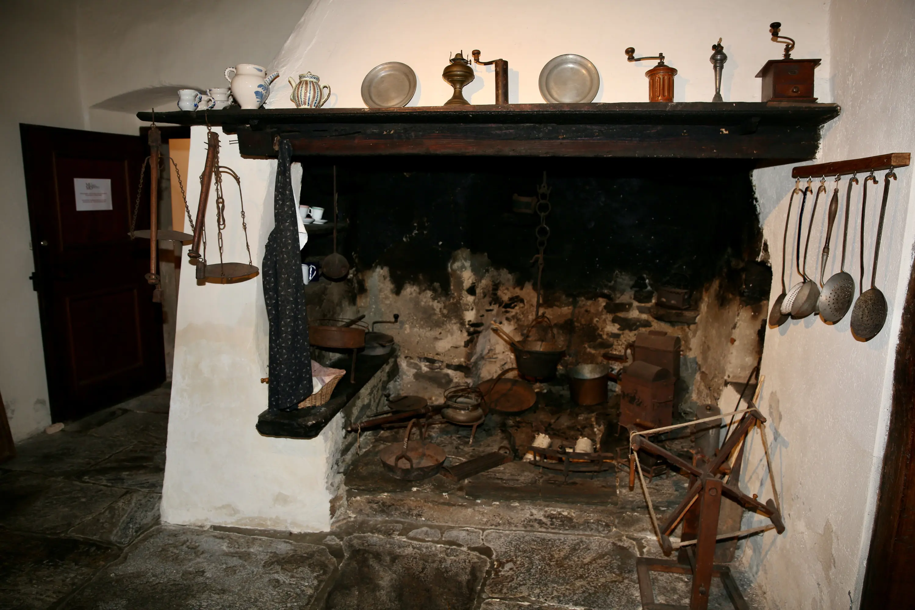 Un'antica cucina con un grande camino in pietra. Sull'asse di legno sopra il camino ci sono vari utensili in metallo e piatti. Appesi alle pareti ci sono mestoli di metallo e altri strumenti da cucina. Il pavimento è in pietra.