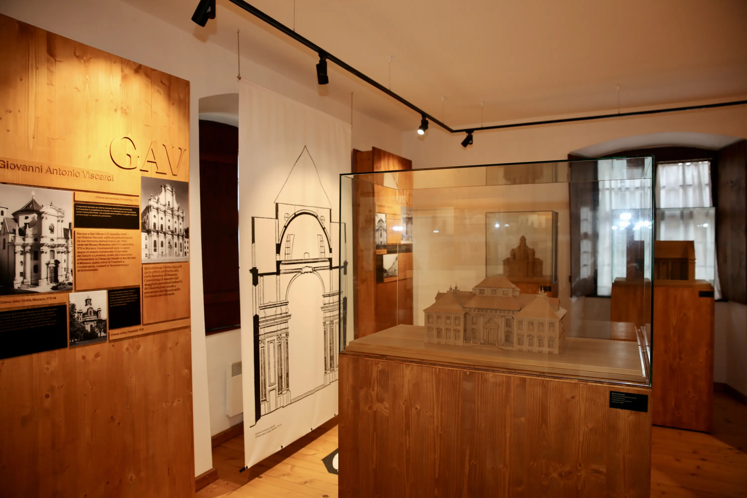 Esposizione all'interno di un museo con pannelli informativi in legno e modelli architettonici in una teca di vetro. Il design è moderno, con un'illuminazione soffusa e dettagli in legno chiaro.