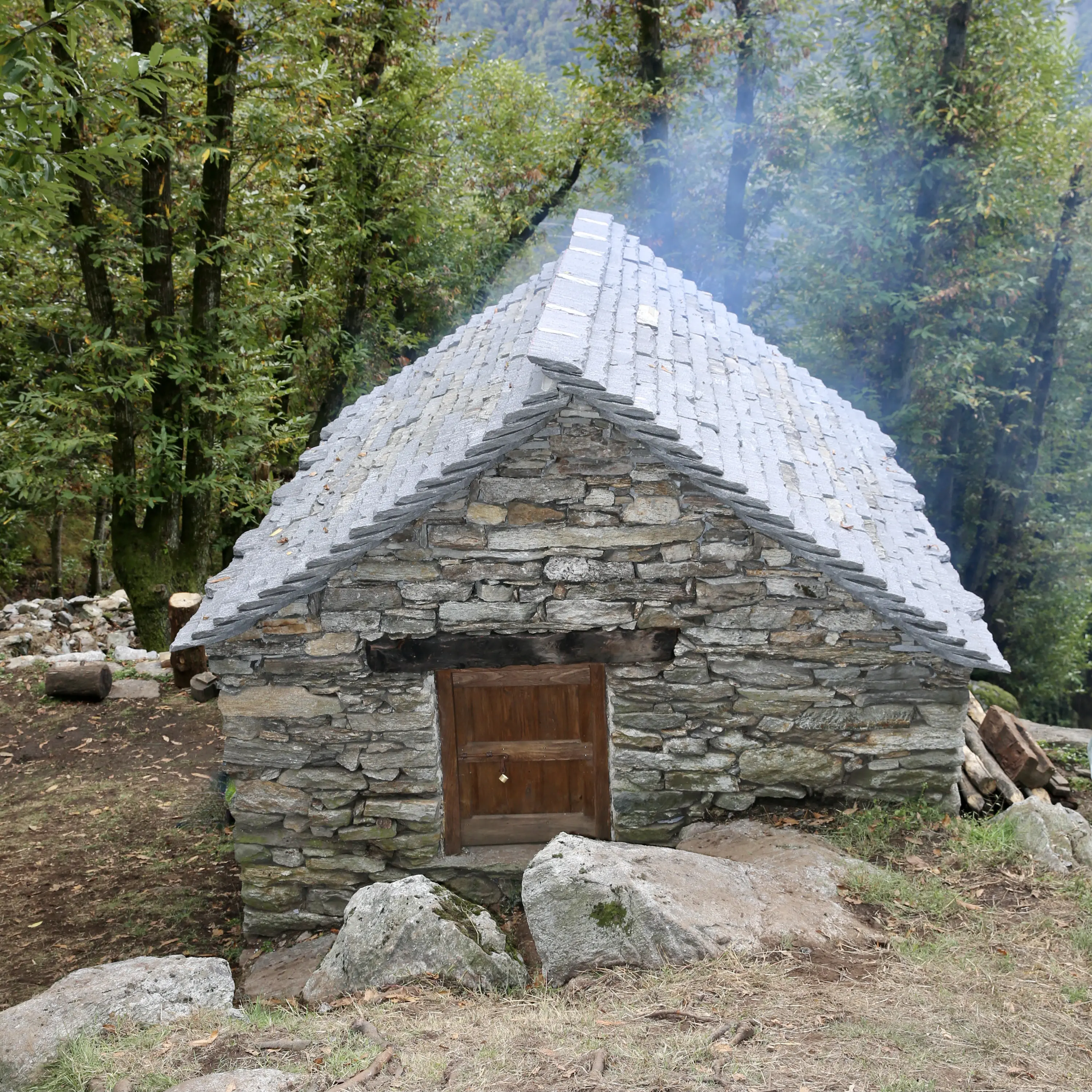 Un piccolo rifugio in pietra con un tetto di lastre, circondato da alberi verdi e un'atmosfera nebbiosa. La porta è di legno, con una superficie rustica.
