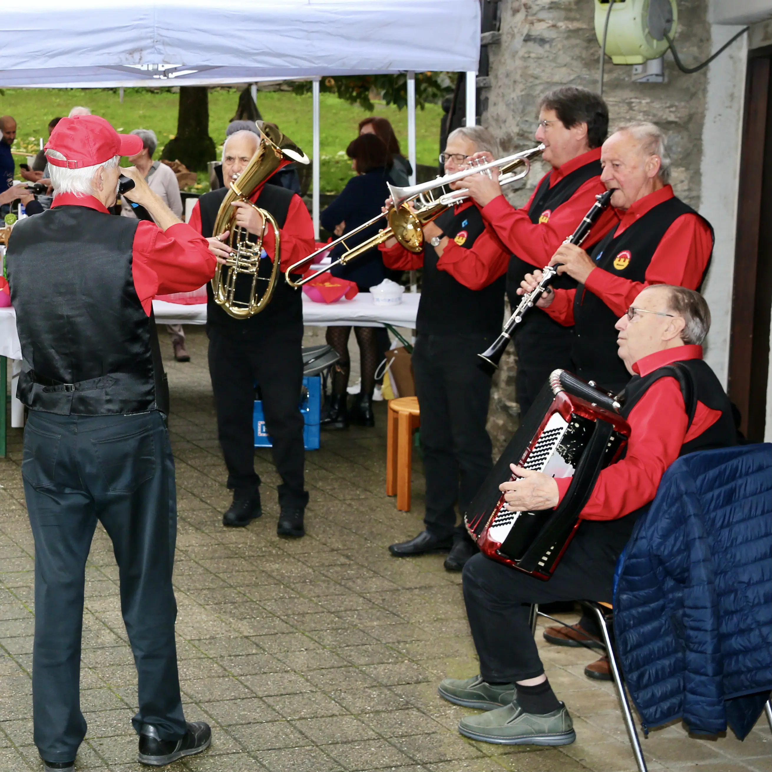 Un gruppo di musicisti in abiti rossi e neri suona strumenti musicali all'aperto. Un uomo anziano suona il trombone, mentre gli altri suonano la tromba, il clarinetto e un fisarmonica. Si possono vedere tendoni e persone sedute in background.