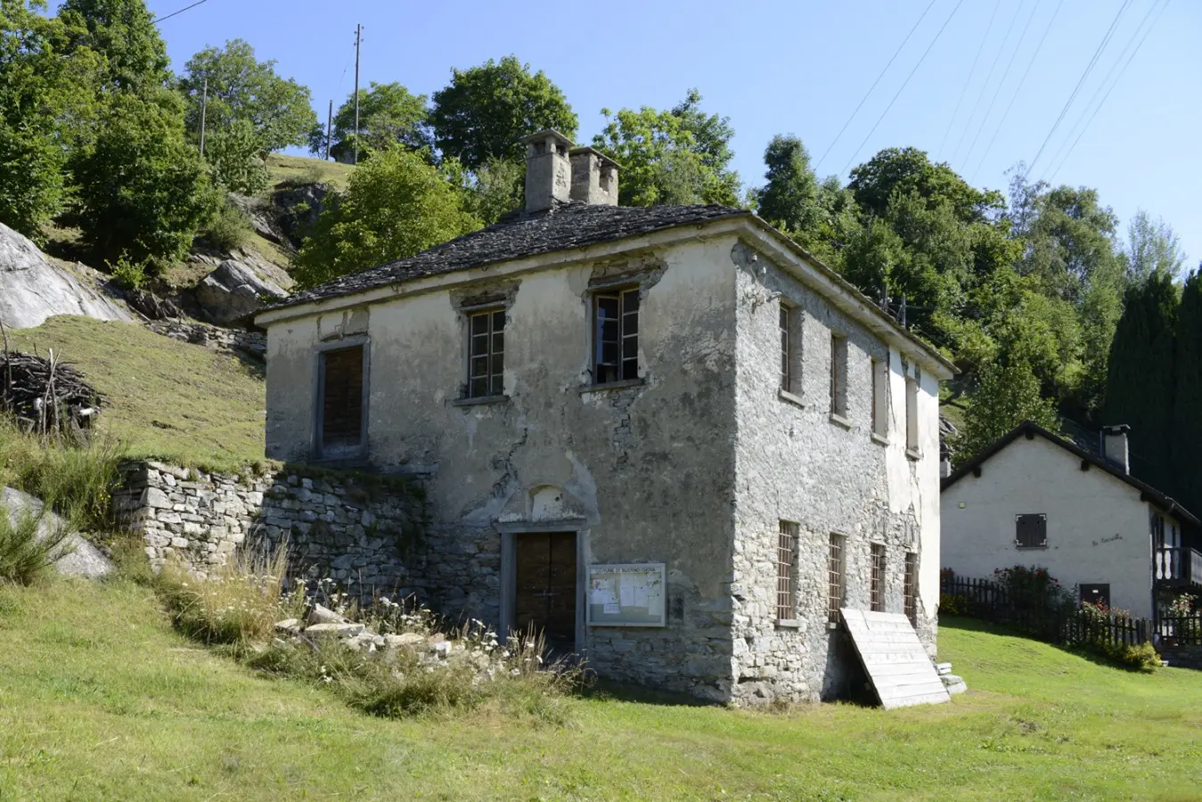 Casa in pietra con pareti grigie e finestre chiuse, circondata da alberi e vegetazione. L'edificio ha un tetto in scandole e una scalinata che conduce all'ingresso. Sullo sfondo, si intravede un'altra costruzione.