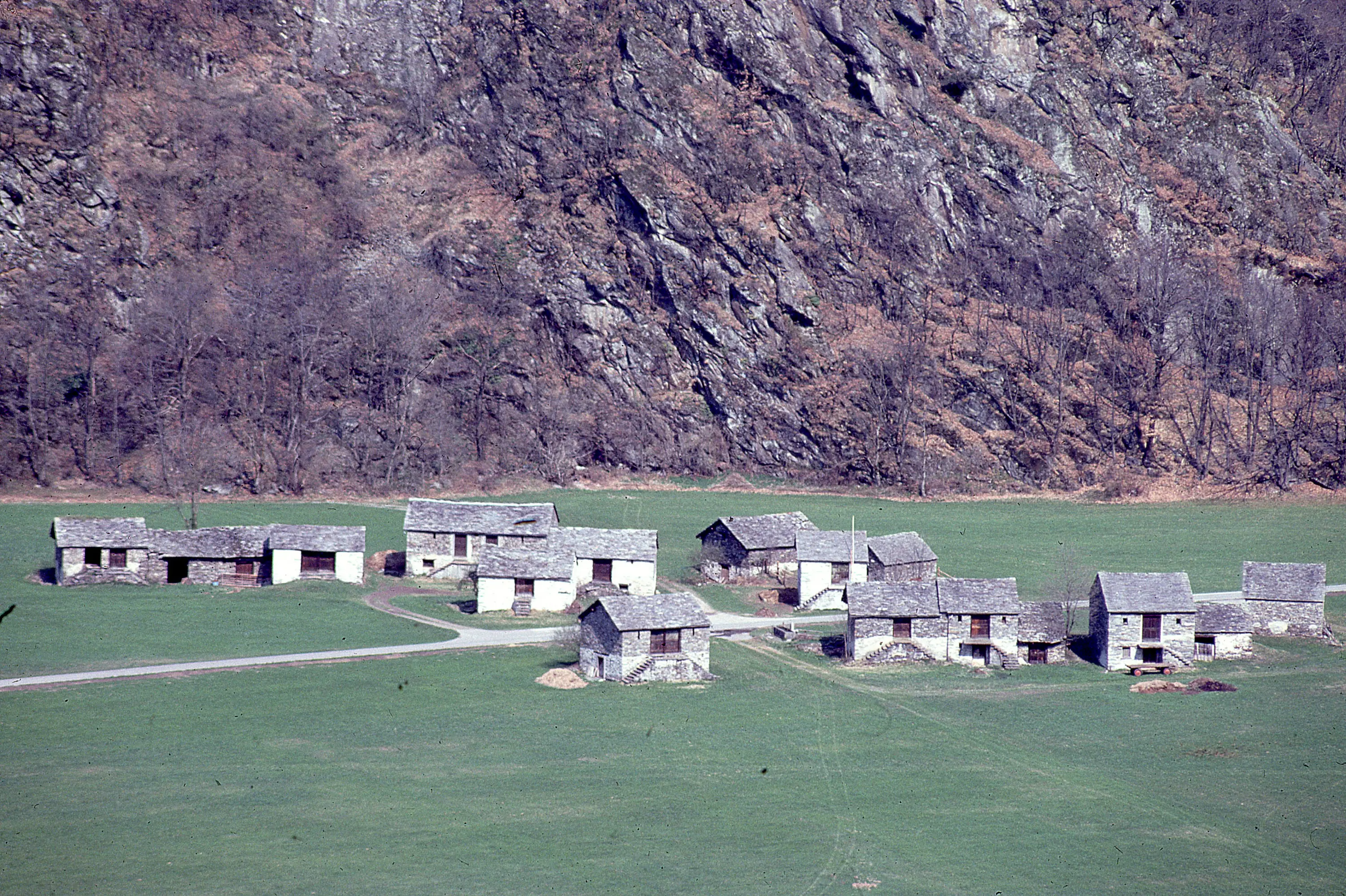 Un gruppo di case in pietra con tetti di legno situate in una valle verde, circondato da un terreno montagnoso e alberi spogli.