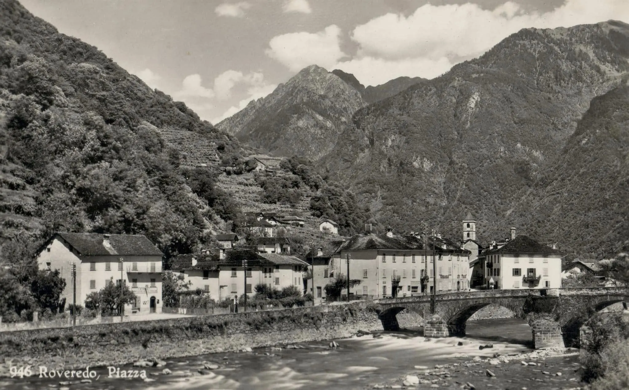 Panorama di Roveredo, con case storiche affacciate sul fiume e montagne sullo sfondo. Un ponte in pietra collega i due lati della piazza.