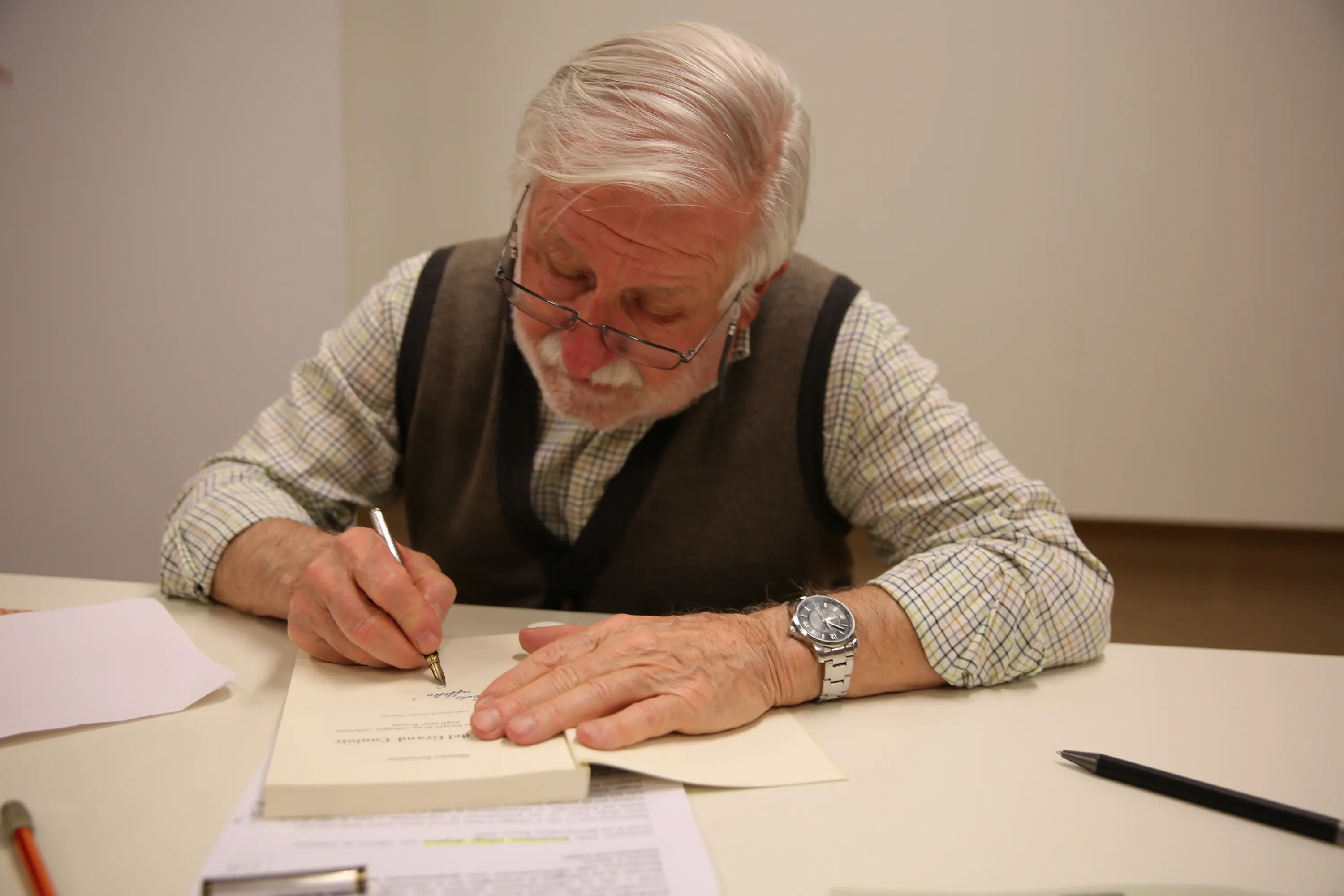 Uomo anziano con capelli bianchi e occhiali, mentre scrive con una penna su un libro aperto. Indossa un gilet sopra una camicia a quadri.