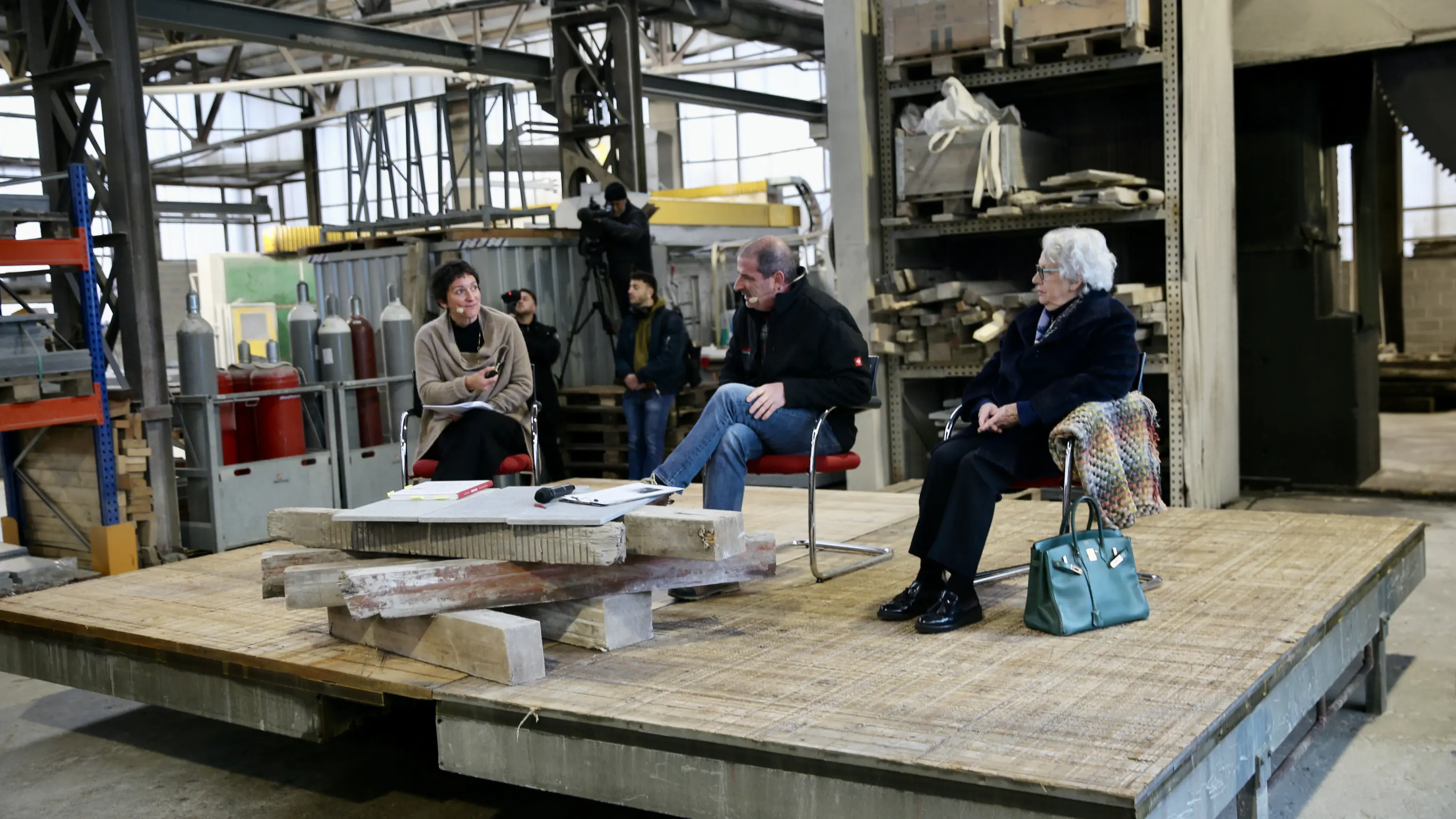 Tre persone sedute su una piattaforma in legno all'interno di un ambiente industriale. Due donne, una in abito scuro e l'altra con una giacca colorata, ascoltano un uomo che parla. Sullo sfondo, alcune attrezzature e attori al lavoro.