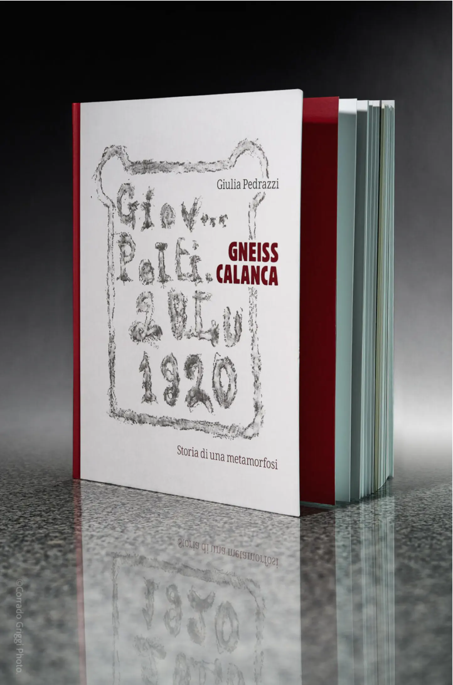 Un libro con copertina bianca e rossa, intitolato "Gneiss Calanca" di Giulia Pedrazzini. La copertina presenta un design semplice con testo grigio e rosso. Sullo sfondo, una superficie scura mette in risalto il libro.