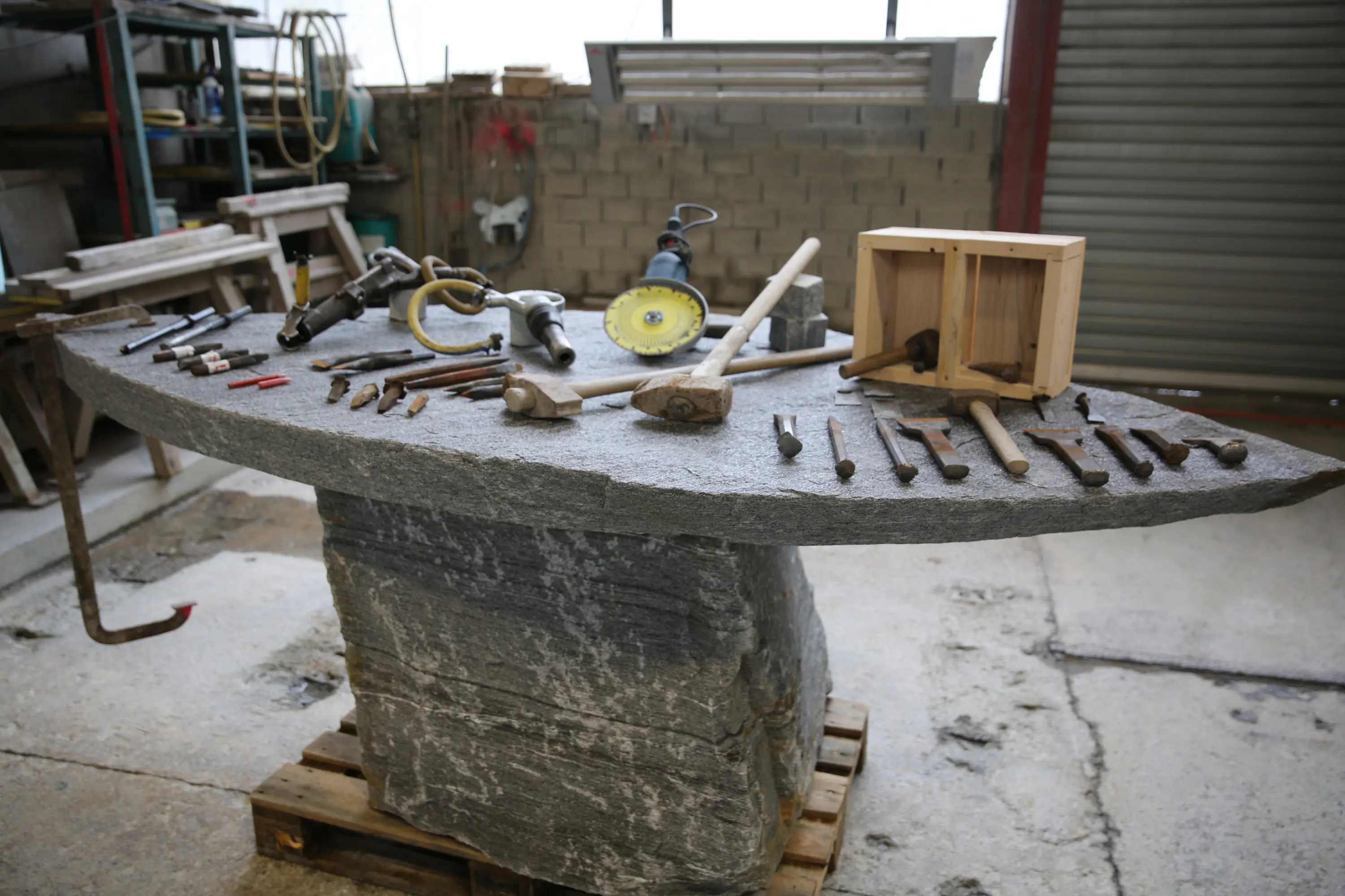 Una tavola di lavoro in pietra grigia con vari attrezzi disposti ordinatamente, tra cui martelli, scalpelli e un trapano. Sullo sfondo si intravedono scaffali e pareti in un ambiente di laboratorio.