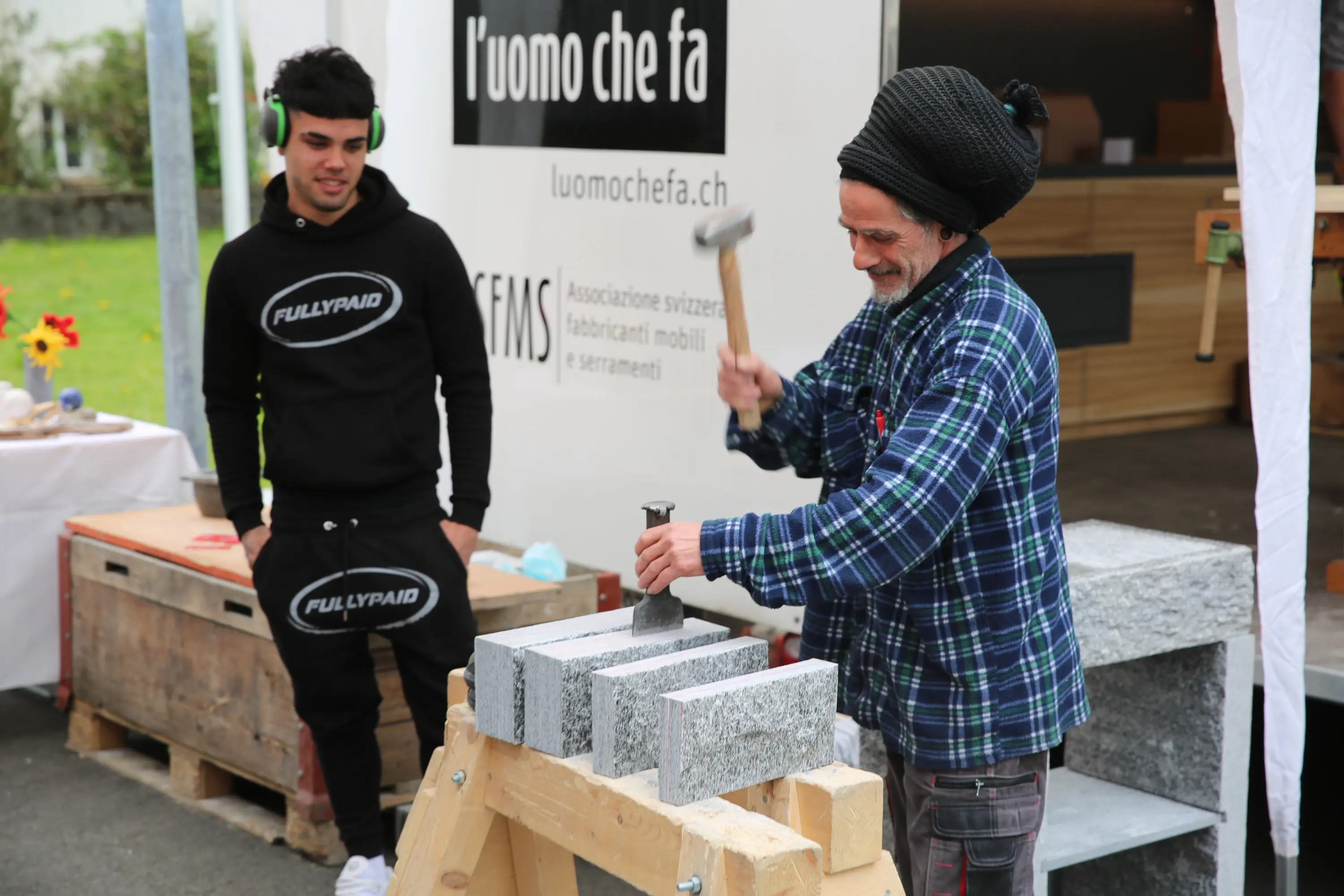 Un artigiano colpisce una pietra con un martello mentre un giovane osserva. Entrambi si trovano in un evento all'aperto, con bancali e uno stand sullo sfondo.