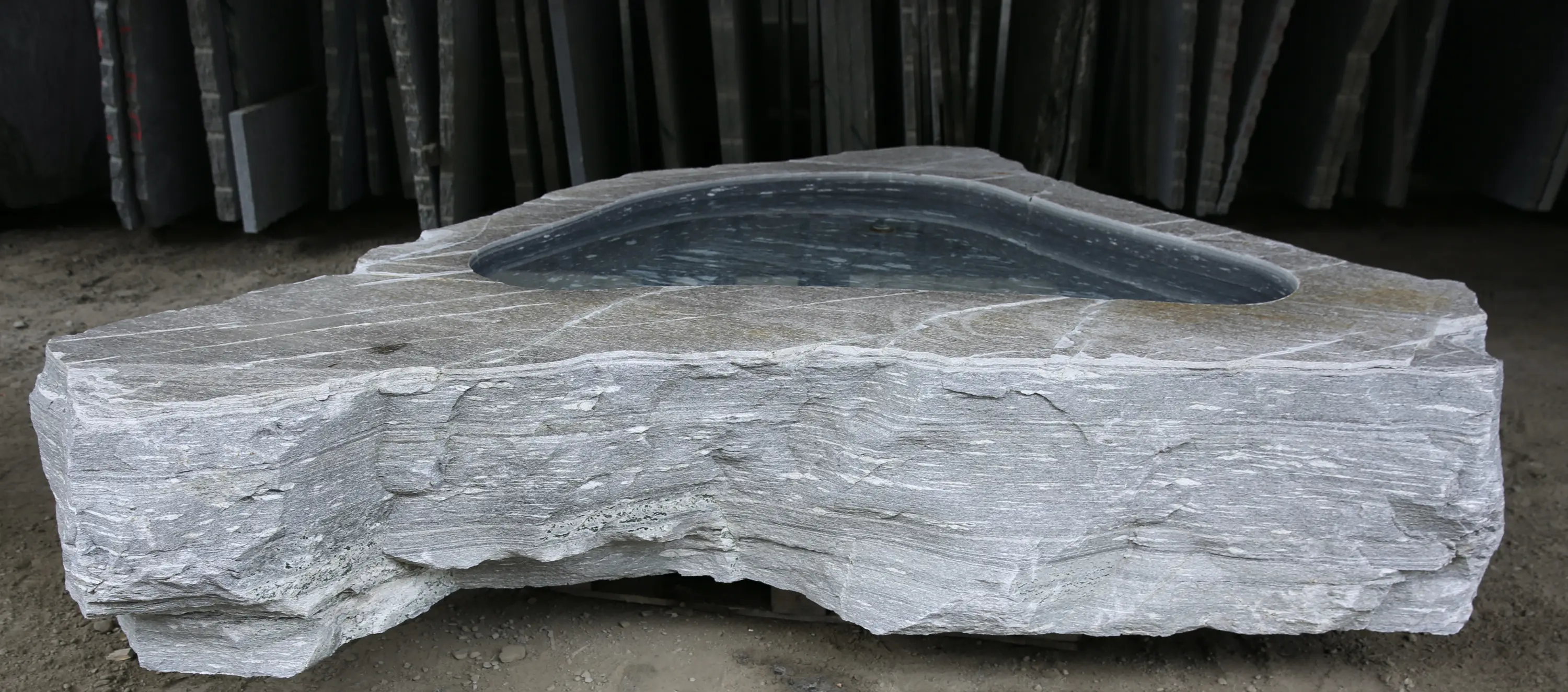 Pietra grigia a forma irregolare con un incavo riempito d'acqua, circondata da lastre di pietra in background.