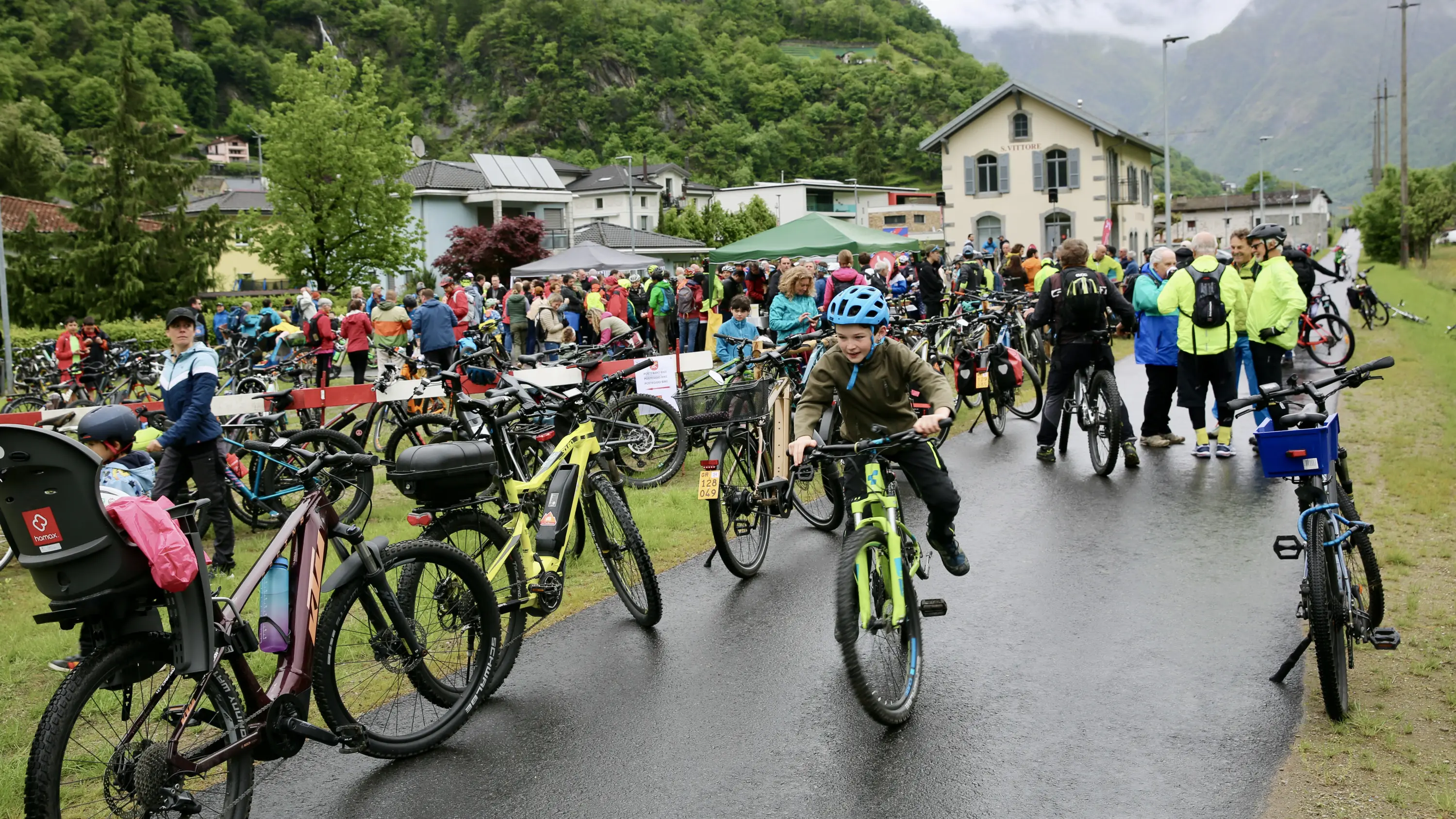 Un gruppo di persone partecipa a un evento ciclistico all'aperto. Alcuni ciclisti stanno montando sulle loro biciclette, mentre altri osservano. Sullo sfondo, si vedono case verdi e alberi. La strada è bagnata, suggerendo che potrebbe aver piovuto.