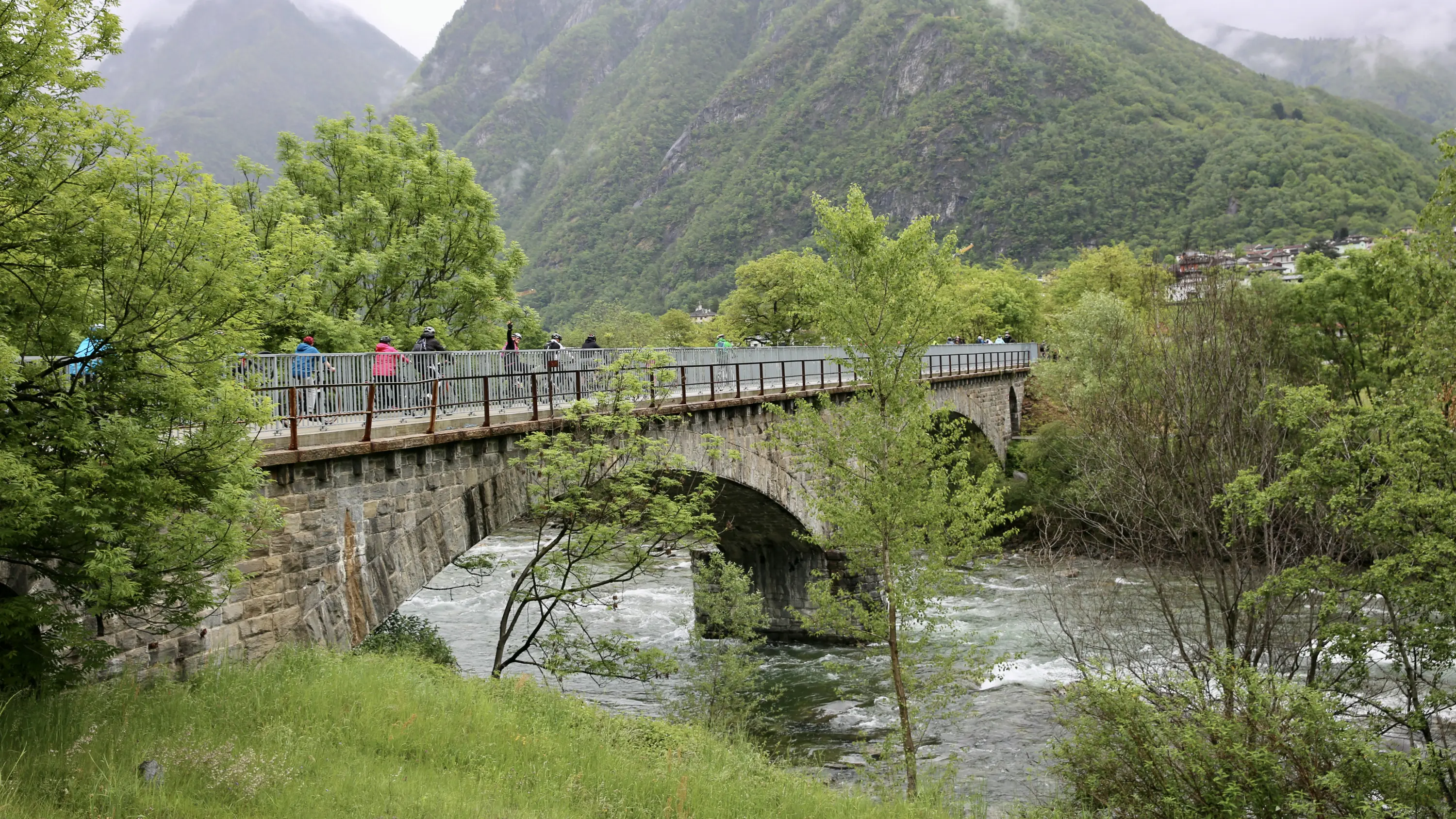 Un ponte in muratura attraversa un fiume con acqua che scorre sotto. La scena è circondata da alberi verdi e colline montuose, con persone che camminano lungo il ponte. Il cielo è nuvoloso, suggerendo un clima umido.