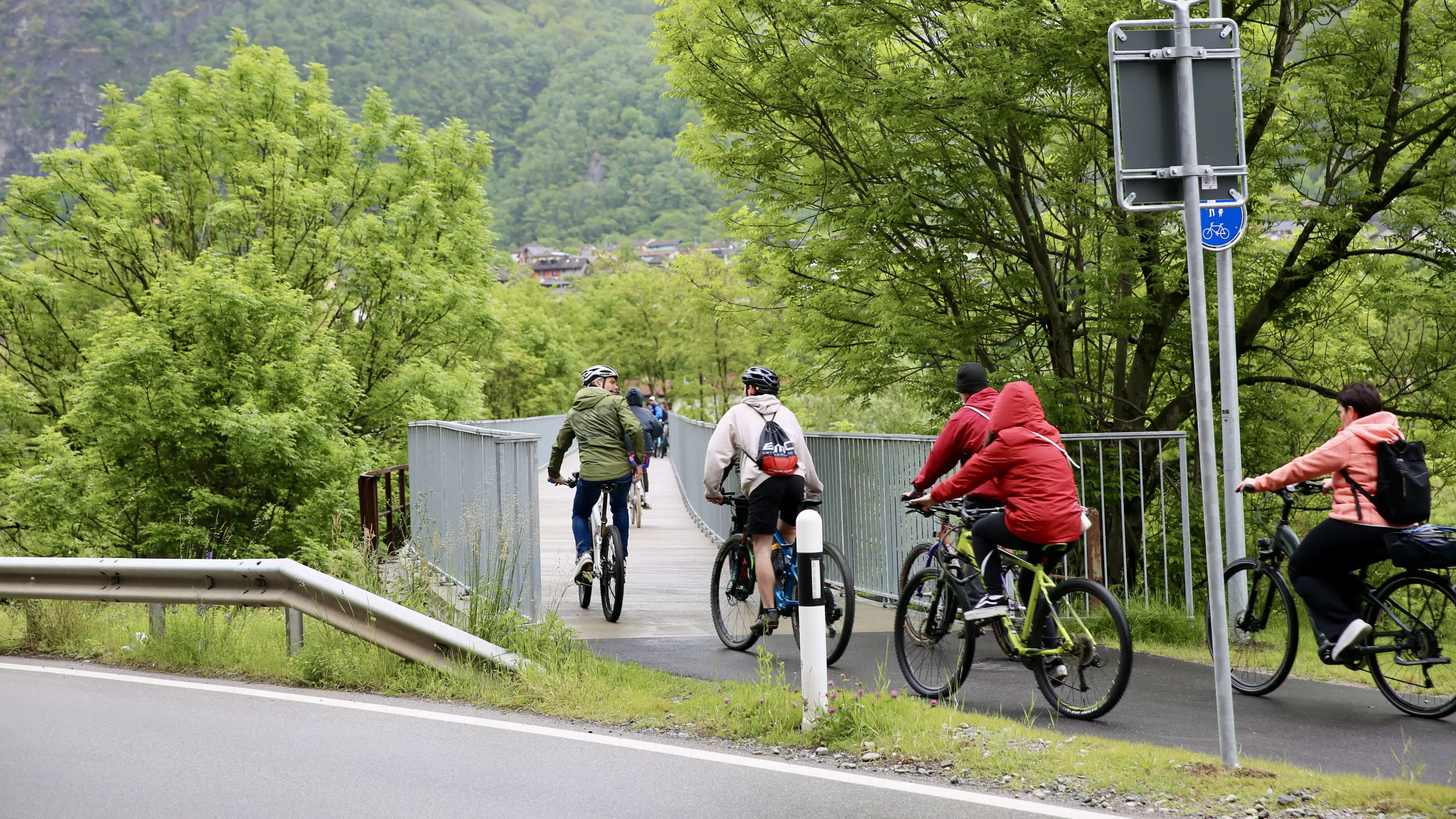 Gruppo di ciclisti che attraversa un ponte in un'area verde, circondati da alberi e colline.