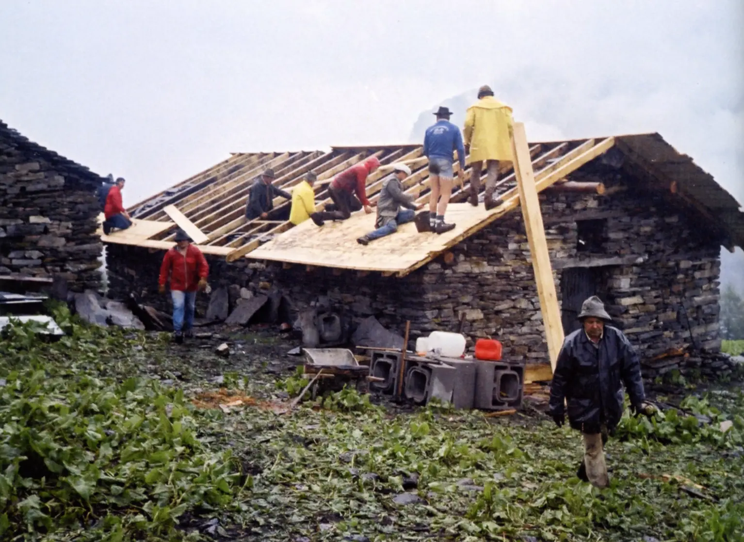Un gruppo di persone lavora sulla riparazione del tetto di una casa in pietra, mentre altre si muovono attorno al cantiere. Il terreno circostante è coperto da foglie verdi e detriti, ed è nuvoloso.