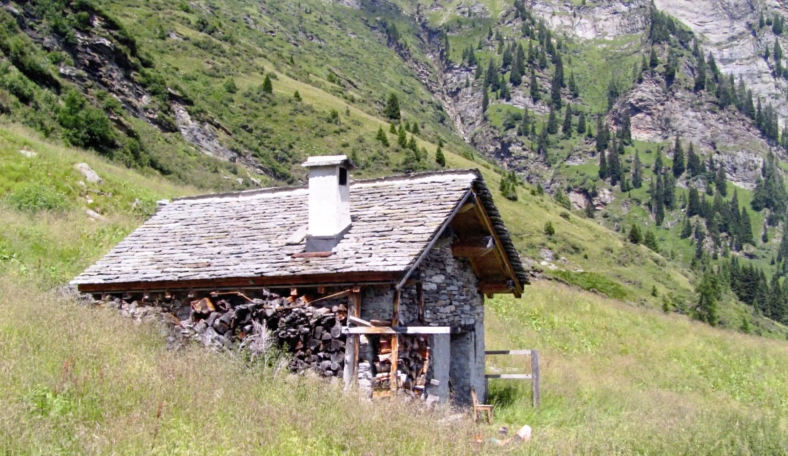 Un caratteristico rifugio in legno e pietra, situato in un'area montuosa verdeggiante. Accanto alla casa ci sono legnaia e un paesaggio di splendide montagne sullo sfondo.