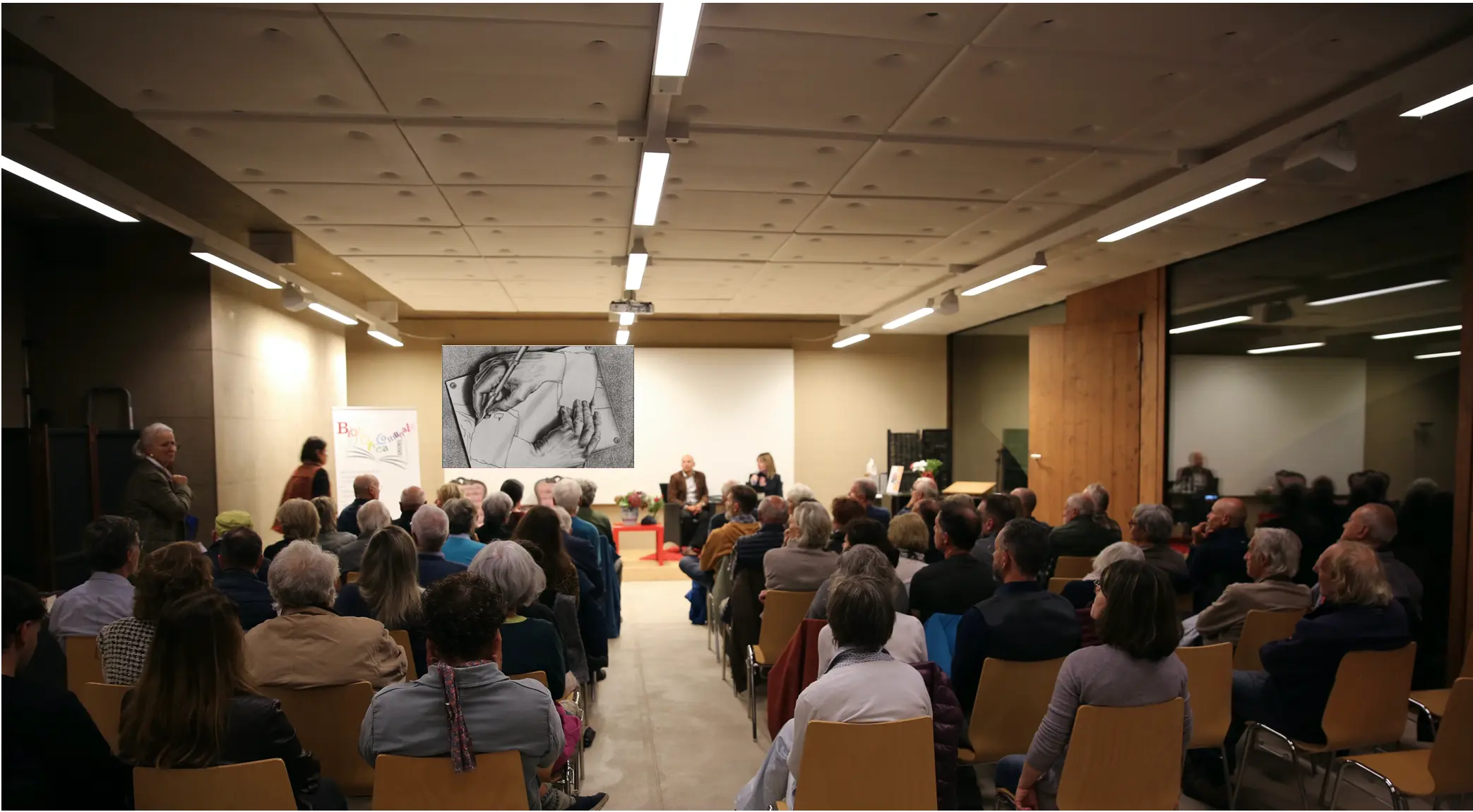 Una sala con molte persone sedute, partecipando a un evento culturale. Sul palco ci sono relatori, mentre sullo sfondo è visibile un'opera d'arte. La scena è ben illuminata, con un'atmosfera conviviale.
