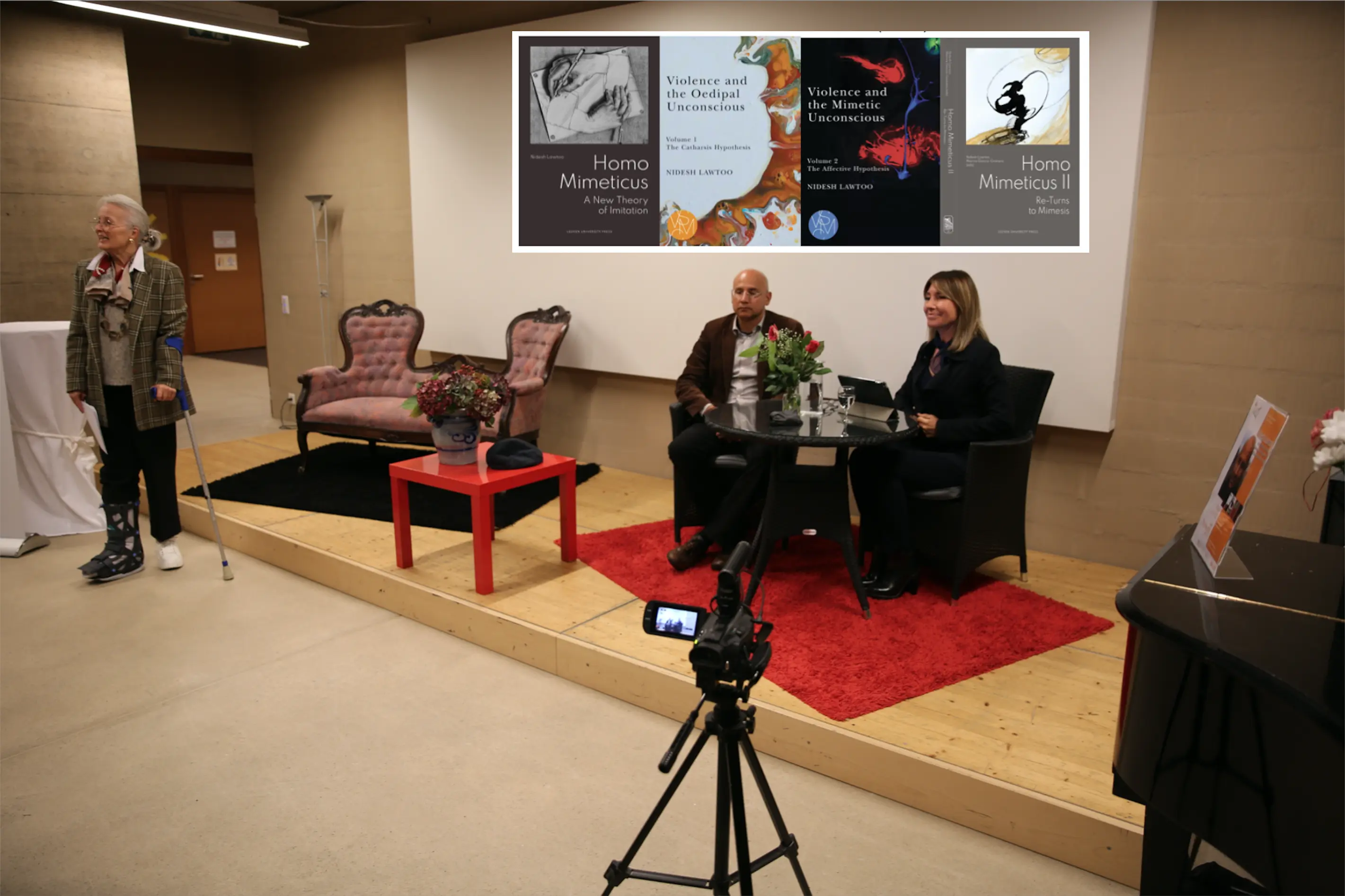 Una presentazione con due relatori seduti a un tavolo, circondati da fiori e libri. Sullo sfondo sono visibili copertine di pubblicazioni. A sinistra, un pubblico partecipa all'evento.