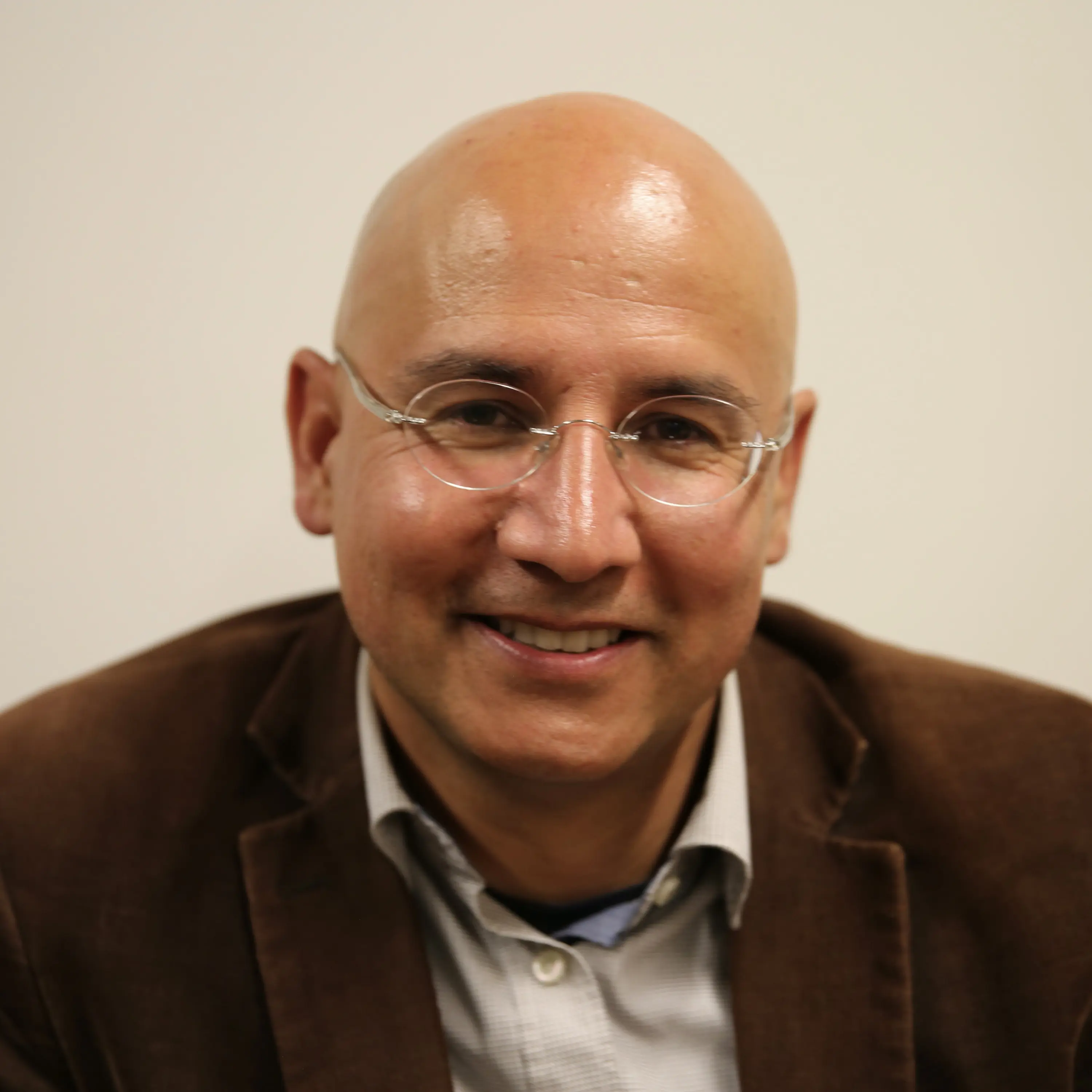 Uomo calvo con occhiali e giacca marrone, sorride. Sfondo neutro.