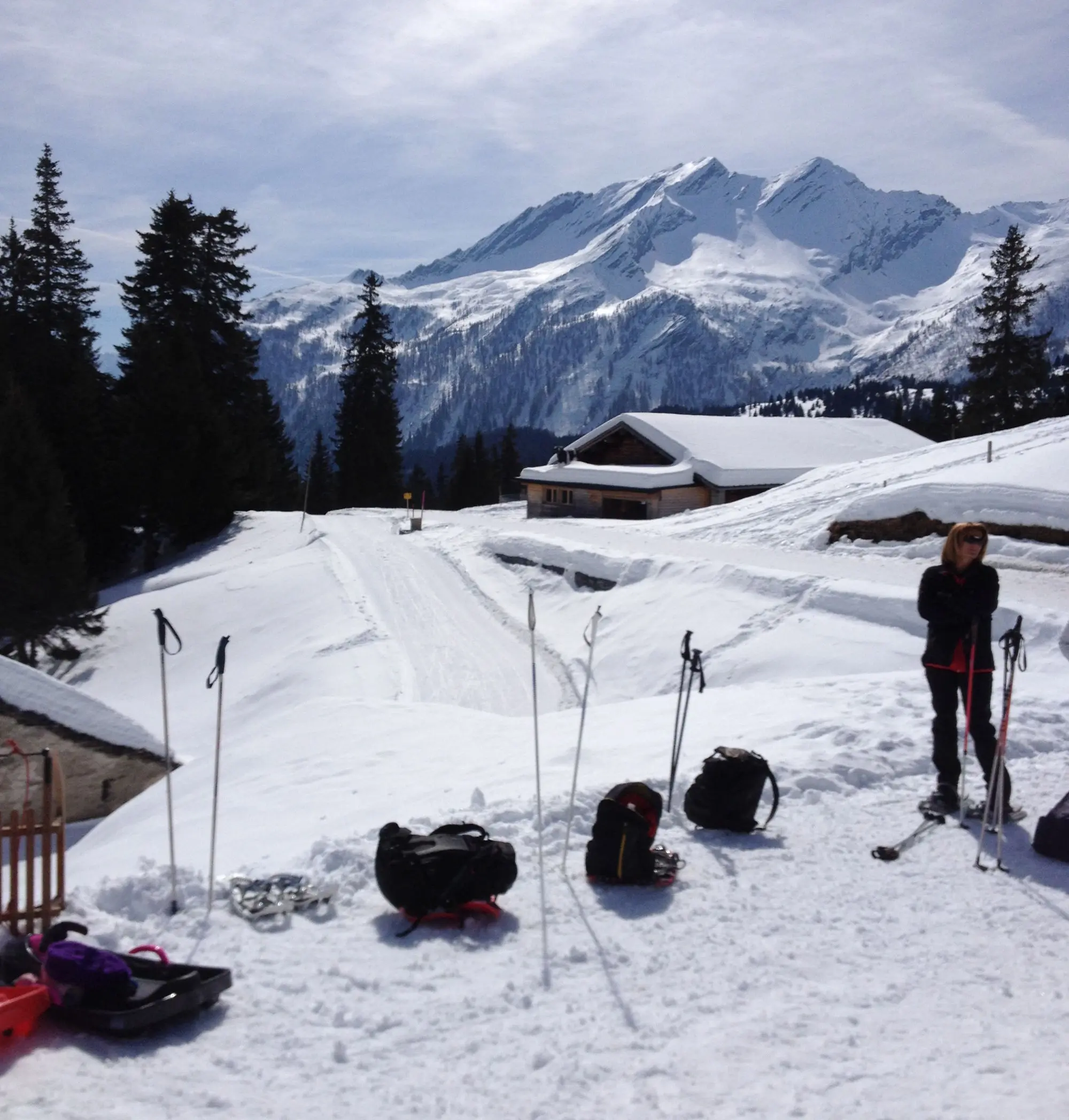 Paesaggio montano innevato con alberi e una casa di legno. Due persone vestite per il freddo si trovano vicino a slitte e attrezzature da sci. Le montagne sullo sfondo sono coperte di neve sotto un cielo parzialmente nuvoloso.