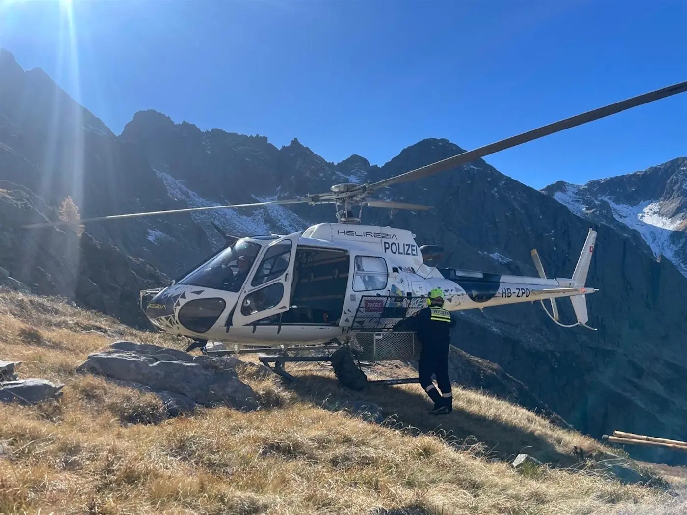 Elicottero della polizia parcheggiato su un terreno erboso e roccioso, con montagne sullo sfondo. Un operatore si avvicina all’elicottero sotto un cielo blu sereno.