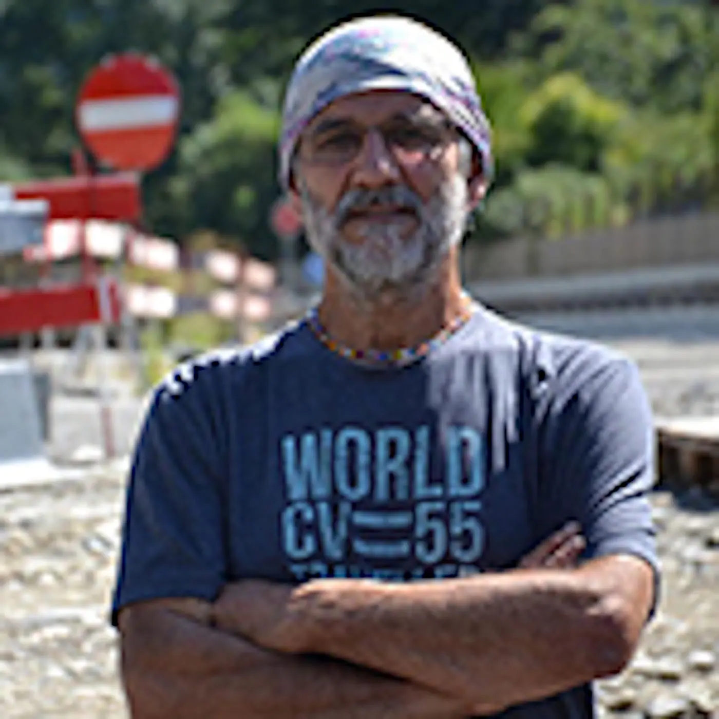 Uomo di mezza età con occhiali e bandana, in piedi con le braccia incrociate, in una zona di costruzione stradale. Sullo sfondo si vedono segnali stradali e cantieri.