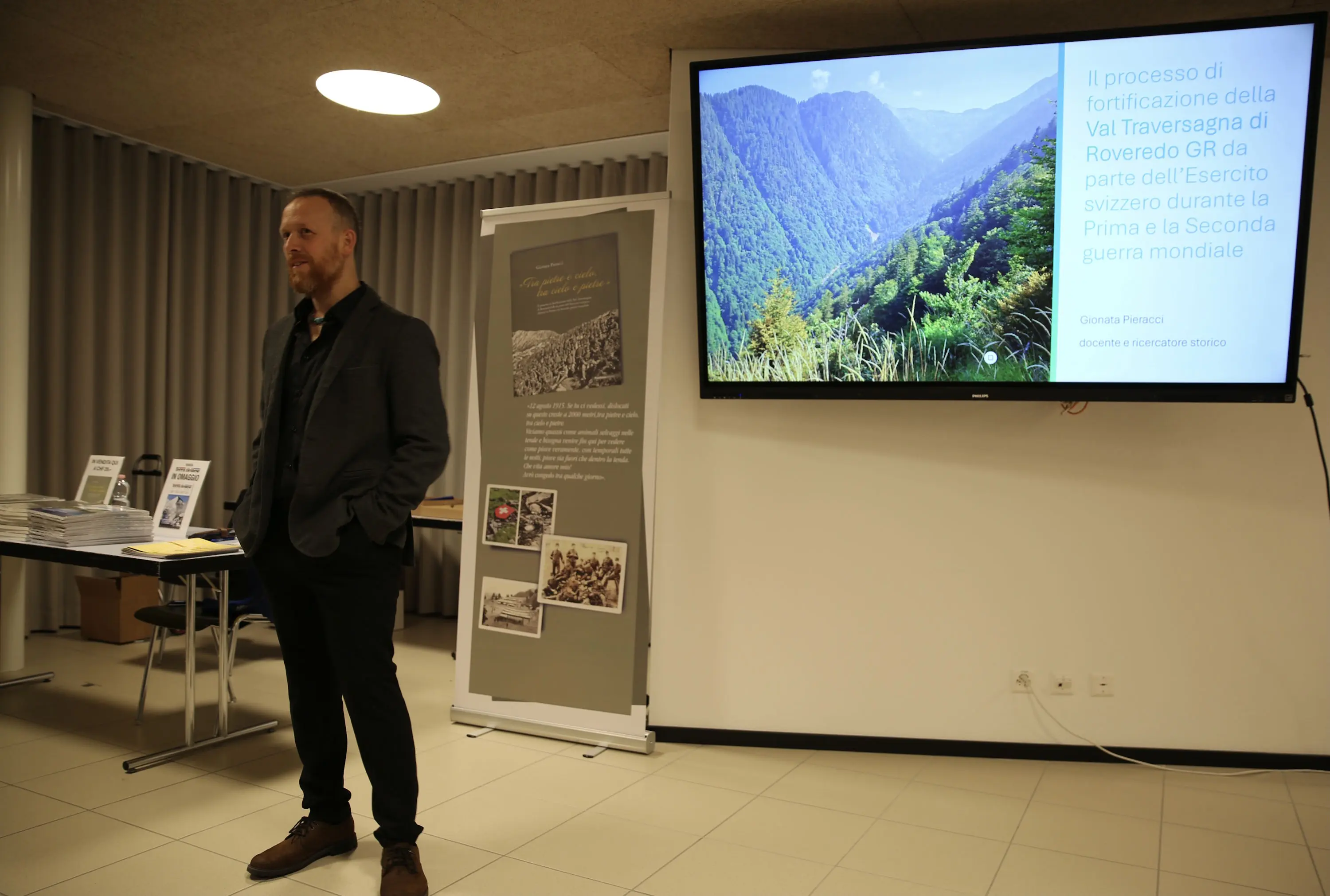 Un relatore in piedi durante una presentazione, con una slide che mostra una vista panoramica della Val Travesagna e il titolo sulla fortificazione durante le guerre mondiali. Sullo sfondo, poster e materiali informativi sono esposti.