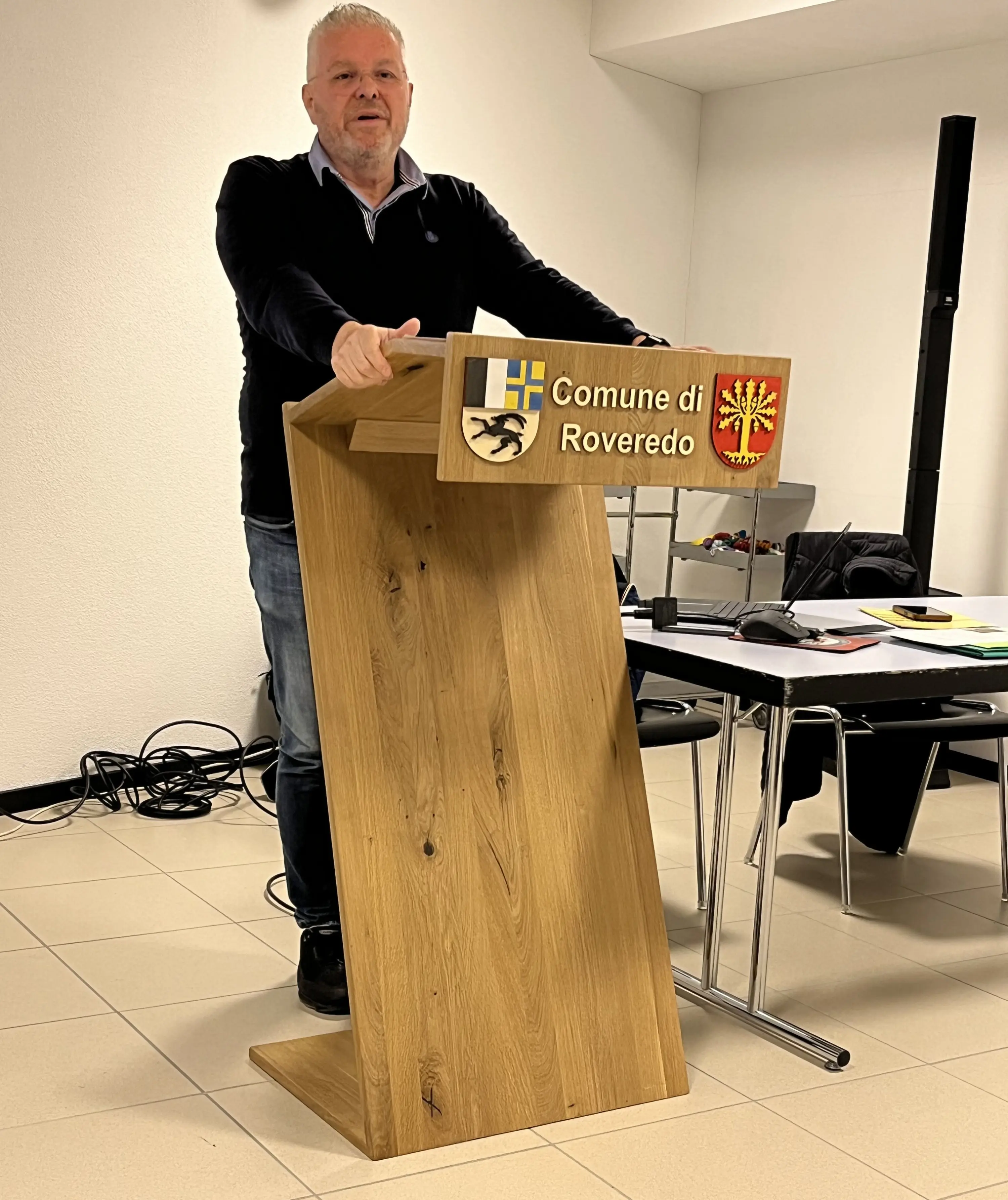 Un uomo in piedi vicino a un podio di legno intagliato con il logo del Comune di Roveredo, in una sala riunioni. Sul tavolo dietro di lui ci sono fogli e attrezzature da ufficio.