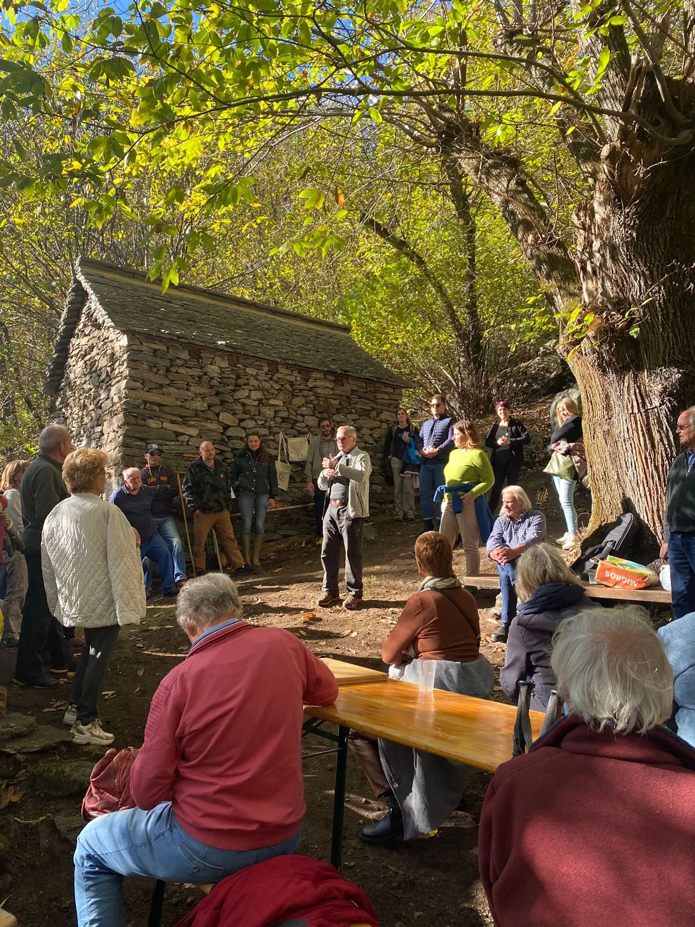 Un gruppo di persone ascolta un oratore di fronte a un vecchio edificio in pietra, circondati da alberi e natura autunnale. Alcuni partecipanti sono seduti a un tavolo, mentre altri si stanze in piedi, attentamente coinvolti nella discussione.