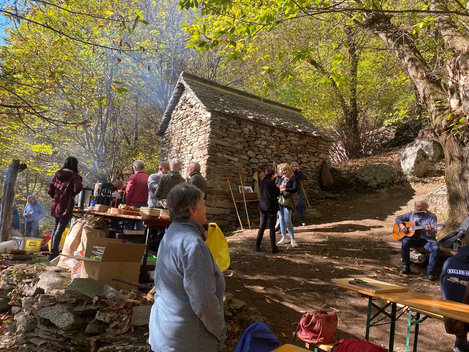 Una festa all'aperto in un bosco, con persone che socializzano vicino a un vecchio edificio in pietra. Alcuni partecipano a un'attività musicale, mentre altri chiacchierano e si godono la natura. Tavoli e scatole sono visibili nel primo piano.