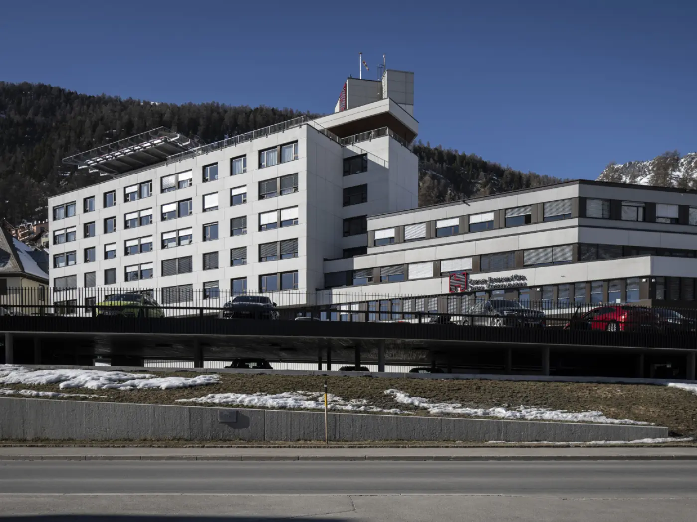 Edificio moderno con facciata bianca e grandi finestre. Sul tetto ci sono pannelli solari. Auto parcheggiate davanti, con una catena montuosa sullo sfondo e un cielo blu sereno.