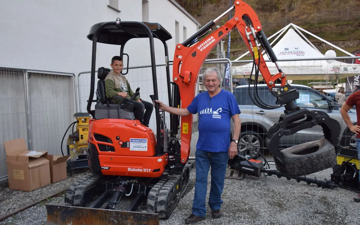 Un uomo adulto sorride accanto a un escavatore rosso, mentre un ragazzo seduto alla guida sembra divertirsi. Sullo sfondo si vedono alcuni veicoli e materiali edili.
