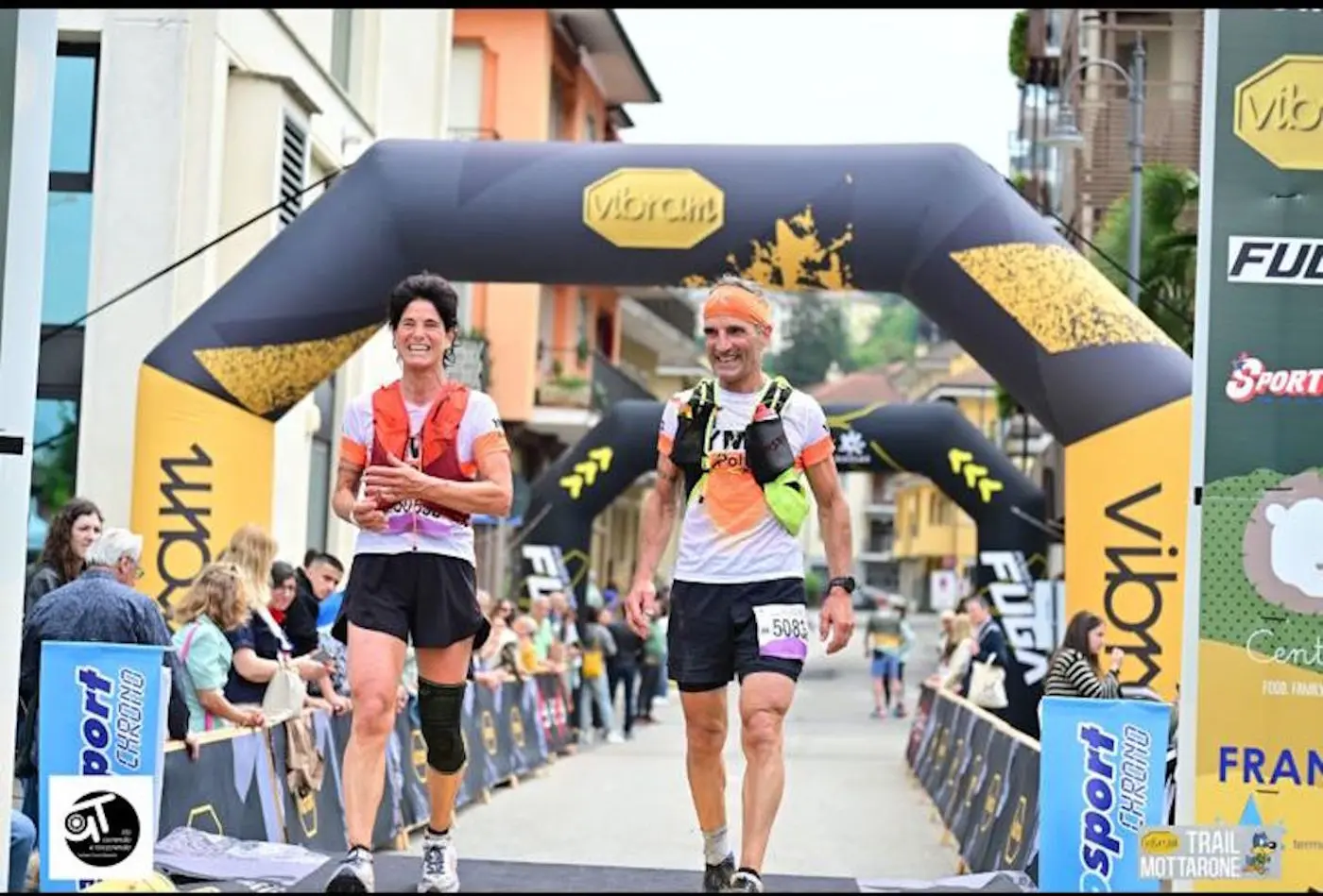 Due corridori sorridenti che tagliano il traguardo di una gara, con un arco gonfiabile sullo sfondo. Diverse persone stanno applaudiando lungo il percorso.
