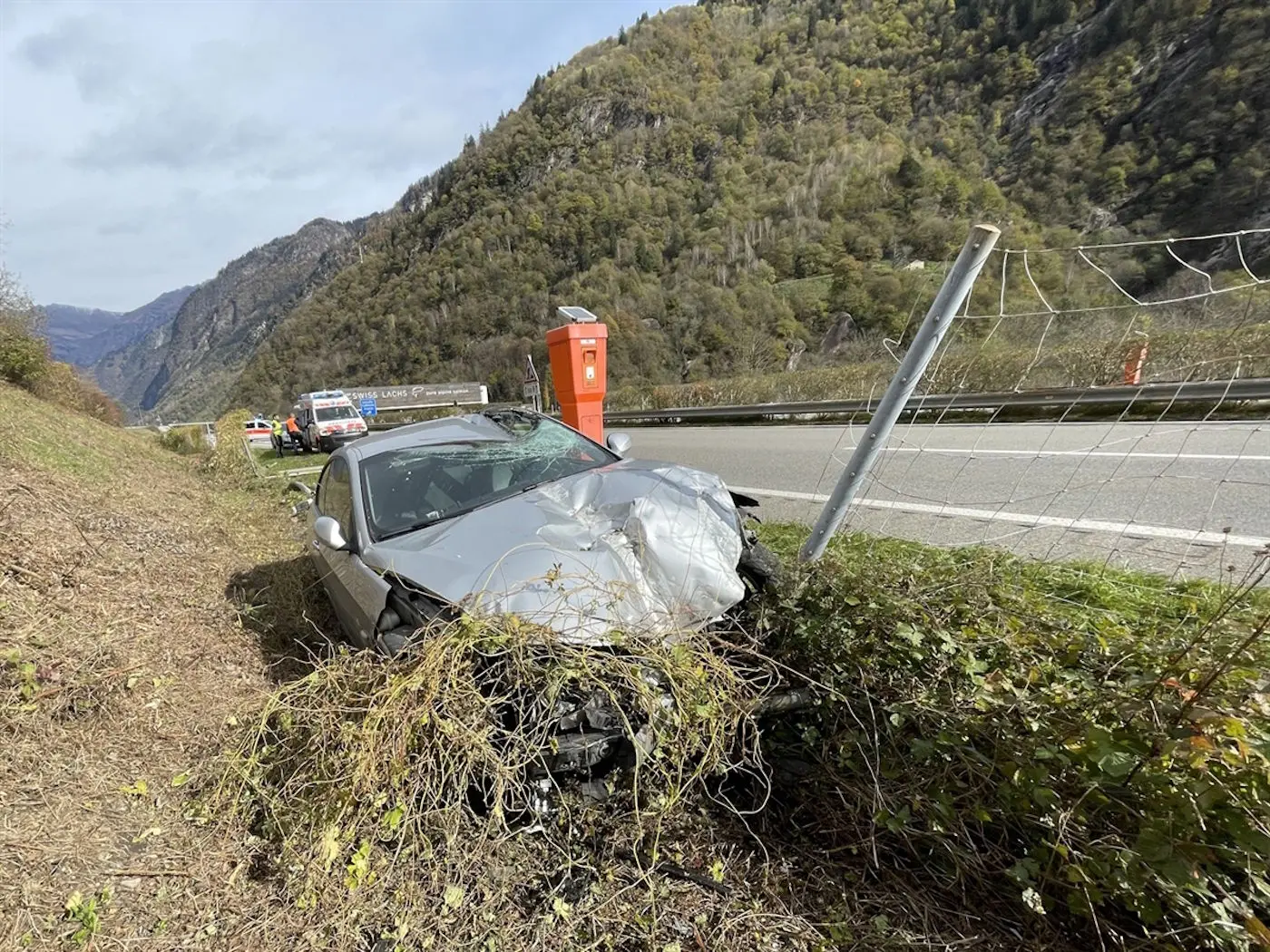 Un'auto grigia danneggiata si trova ai margini di una strada, circondata da arbusti. Sullo sfondo, si vedono delle montagne e un veicolo dei soccorsi. Un box per la raccolta della posta è visibile a lato della strada.