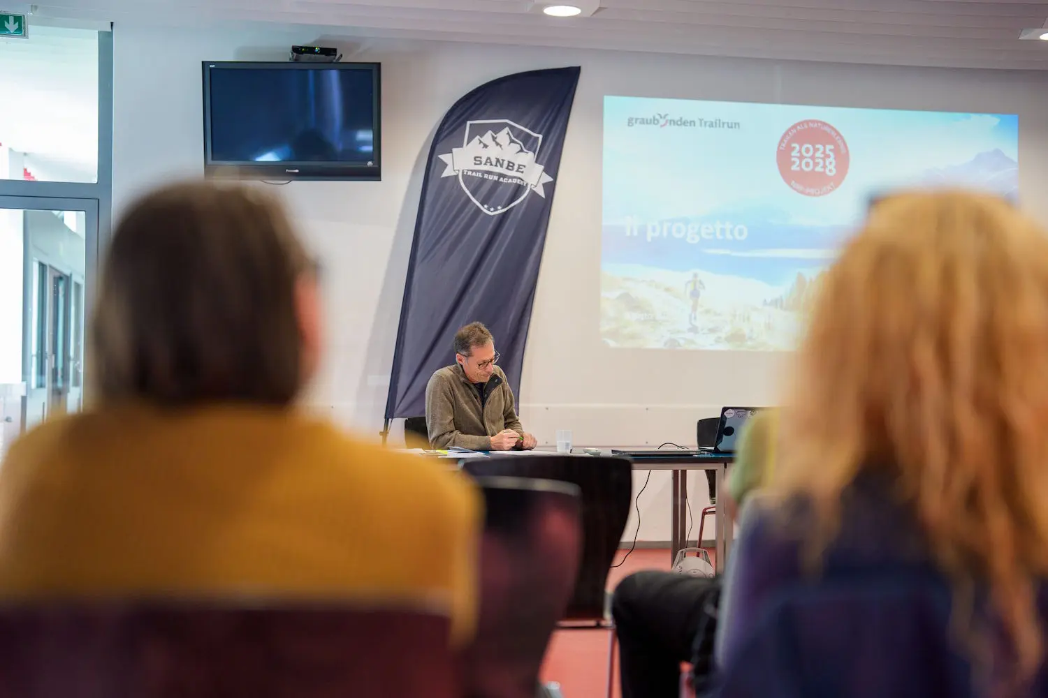 Un uomo seduto a un tavolo presenta un progetto durante una conferenza. Sullo sfondo, si vede uno schermo con informazioni relative al progetto e un banner con il logo "SANBE". La sala è affollata, ma le persone in primo piano sono sfocate.